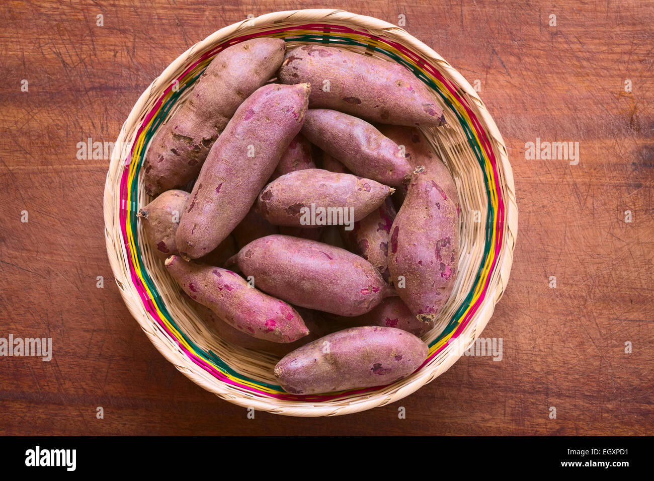 Colpo di overhead di materie viola la patata dolce (lat. Ipomoea batatas) nel cesto tessuto sulla tavola di legno fotografati con luce naturale Foto Stock