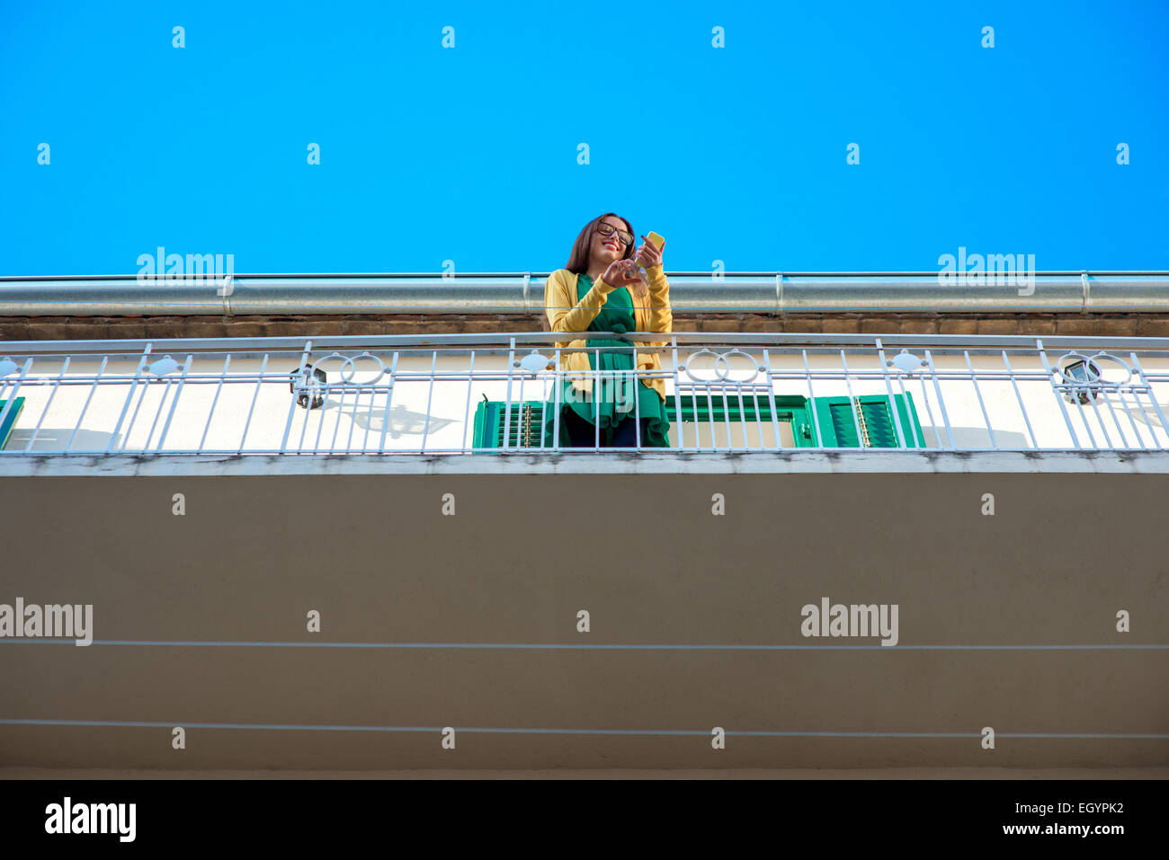 Giovane donna in Felpa gialla e bicchieri utilizzando il telefono cellulare sul balcone con persiane alle finestre nel vecchio edificio. Piano generale. Foto Stock