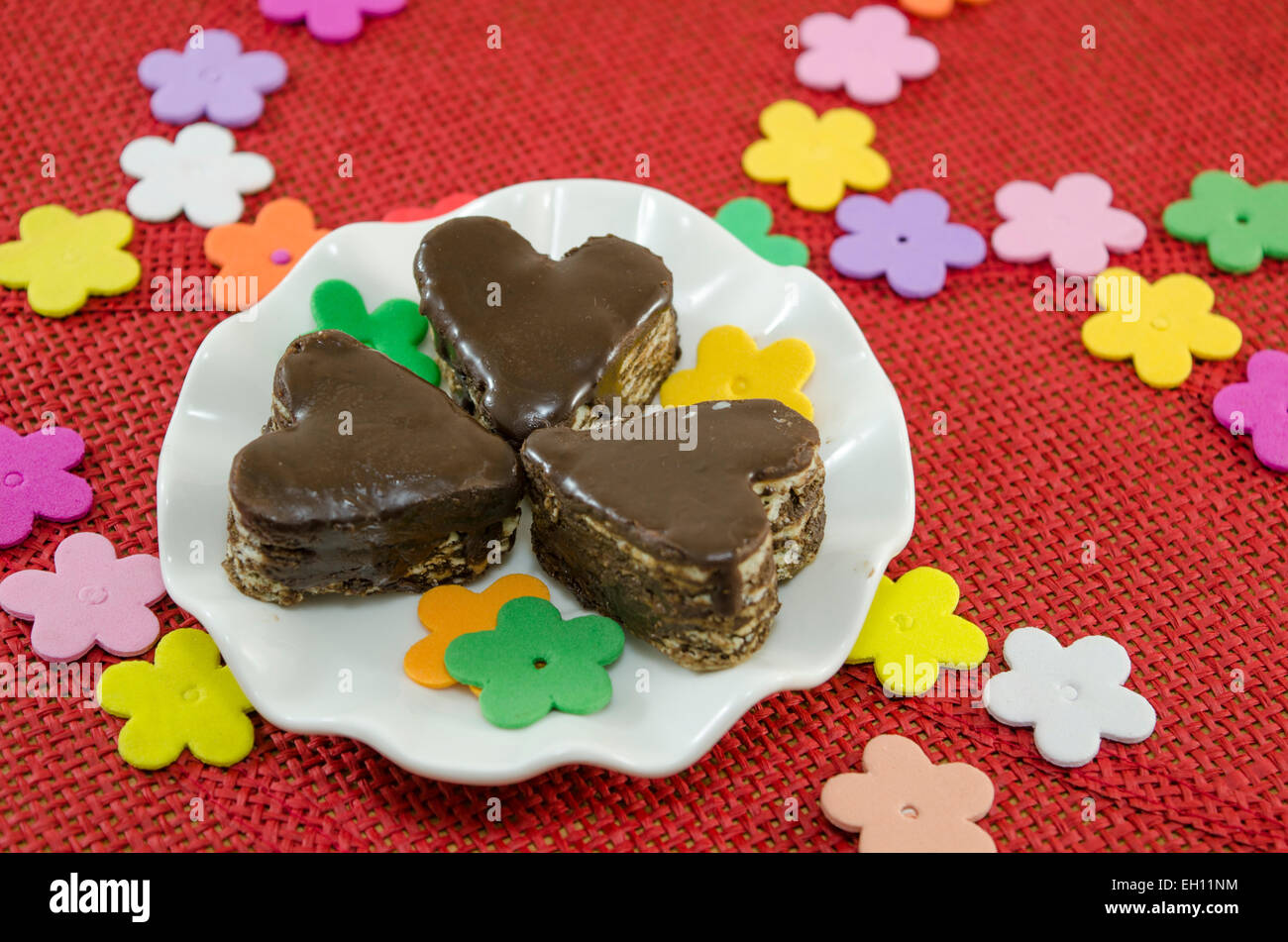 Al cioccolato a forma di cuore i cookie su una piastra decorato con fiori di carta su una tovaglia rosso Foto Stock