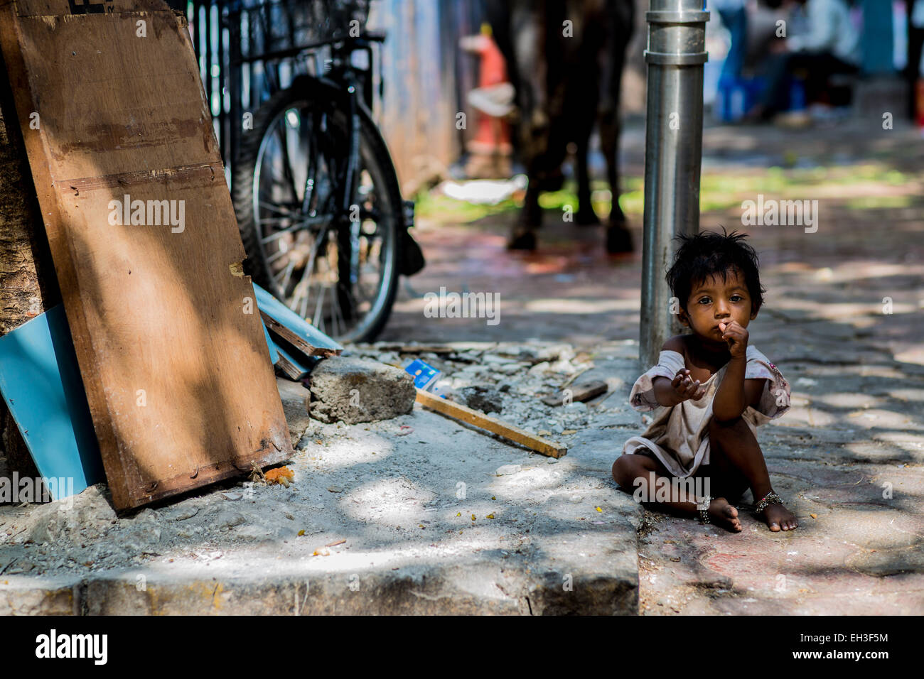 Un piccolo homeless street bambino è seduta sul ciglio della strada guardando la telecamera con la sua mano. Foto Stock