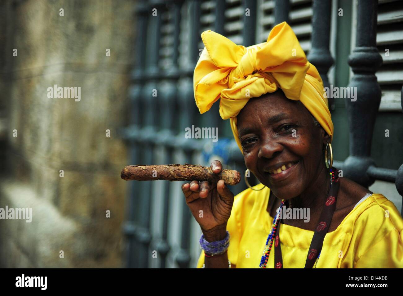 Cuba, La Habana, Nora, senior donna vestita con il tradizionale abito giallo di fumare il sigaro Foto Stock