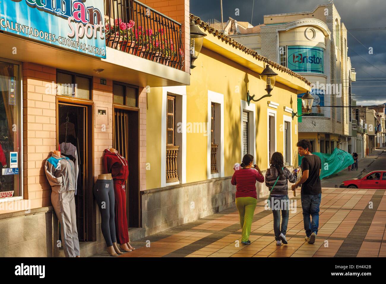 Ecuador, Imbabura, Atuntaqui, scena di strada di persone che passano di fronte a un negozio di abbigliamento sul cielo tempestoso al tramonto Foto Stock