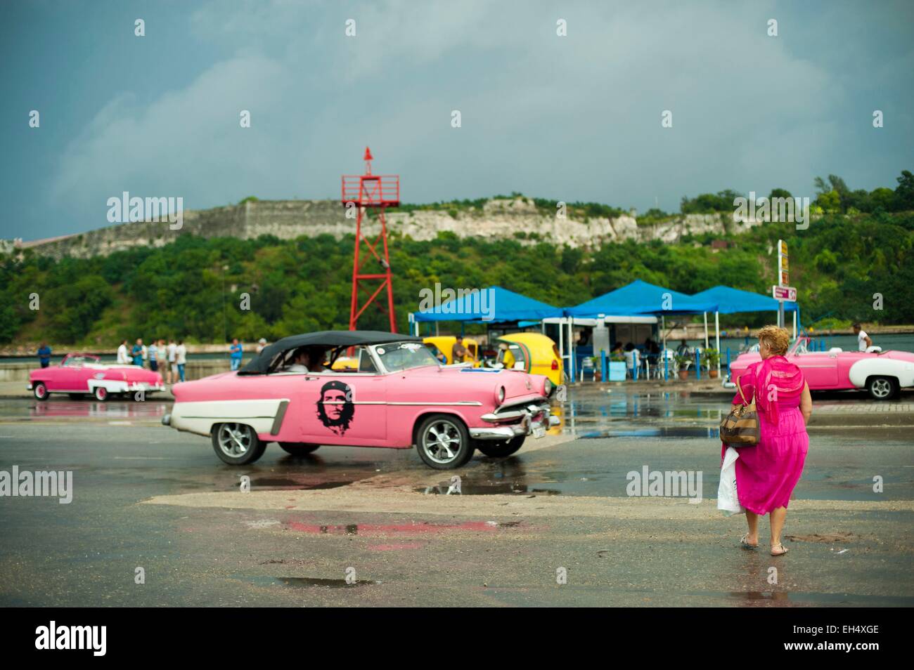 Cuba, La Habana, donna in abito rosa attraversando la strada con molti vecchi american chevrolet parcheggiata Foto Stock
