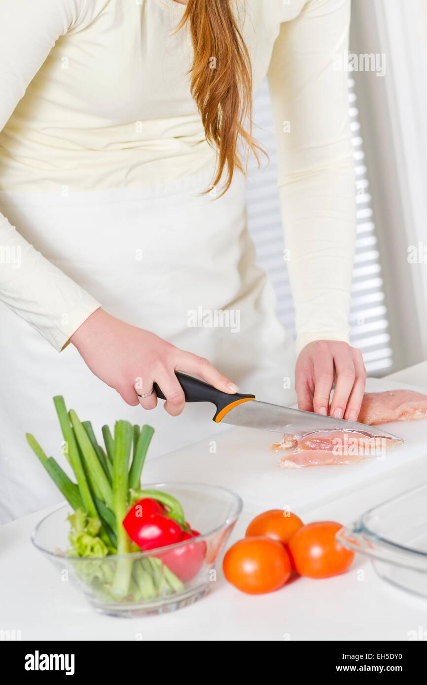 Giovane donna in una cucina bianca tagliare la carne di pollo filetto. Ortaggio sul banco di lavoro. Foto Stock