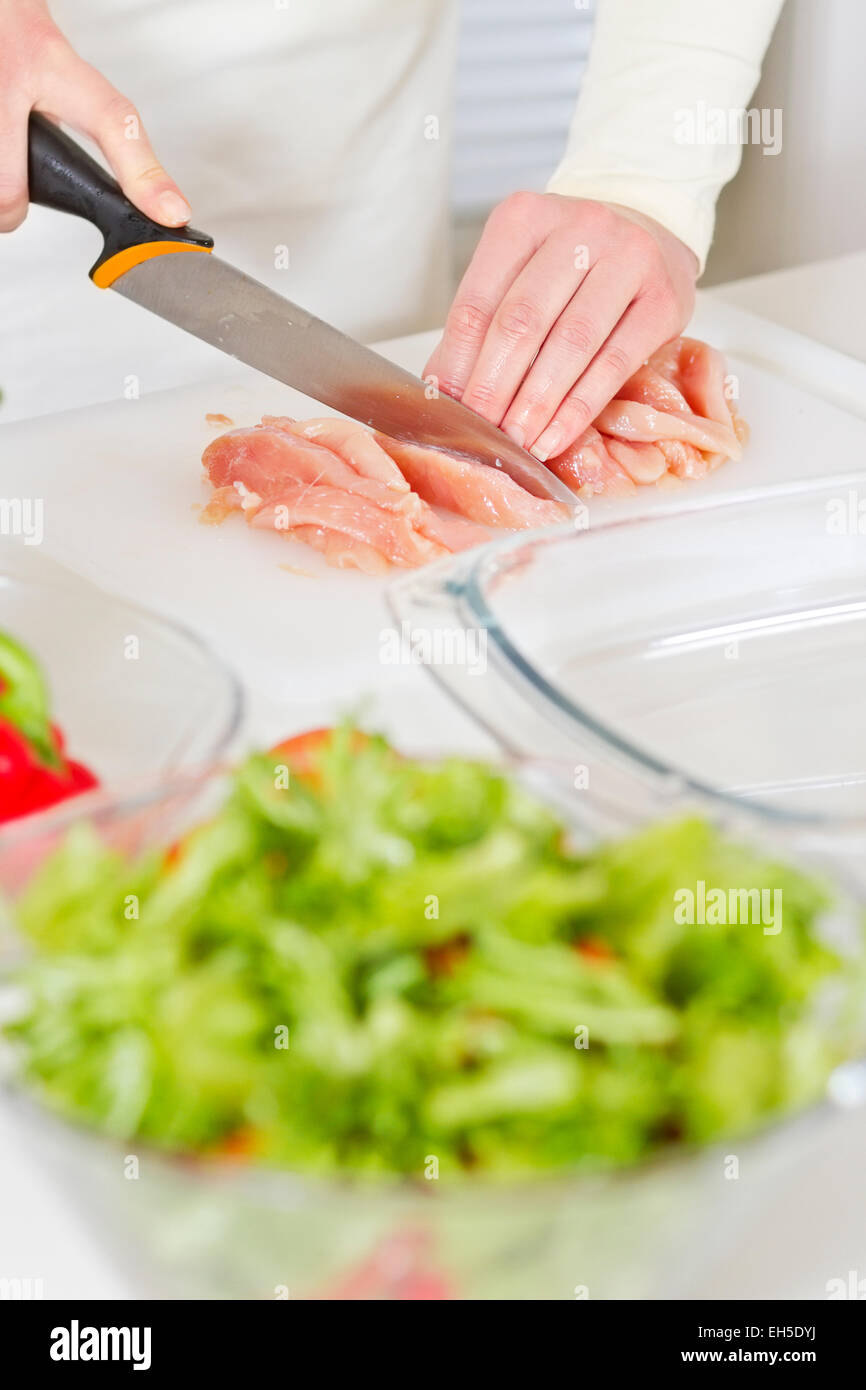 Giovane donna in una cucina bianca tagliare la carne di pollo filetto. Verdure. Foto Stock
