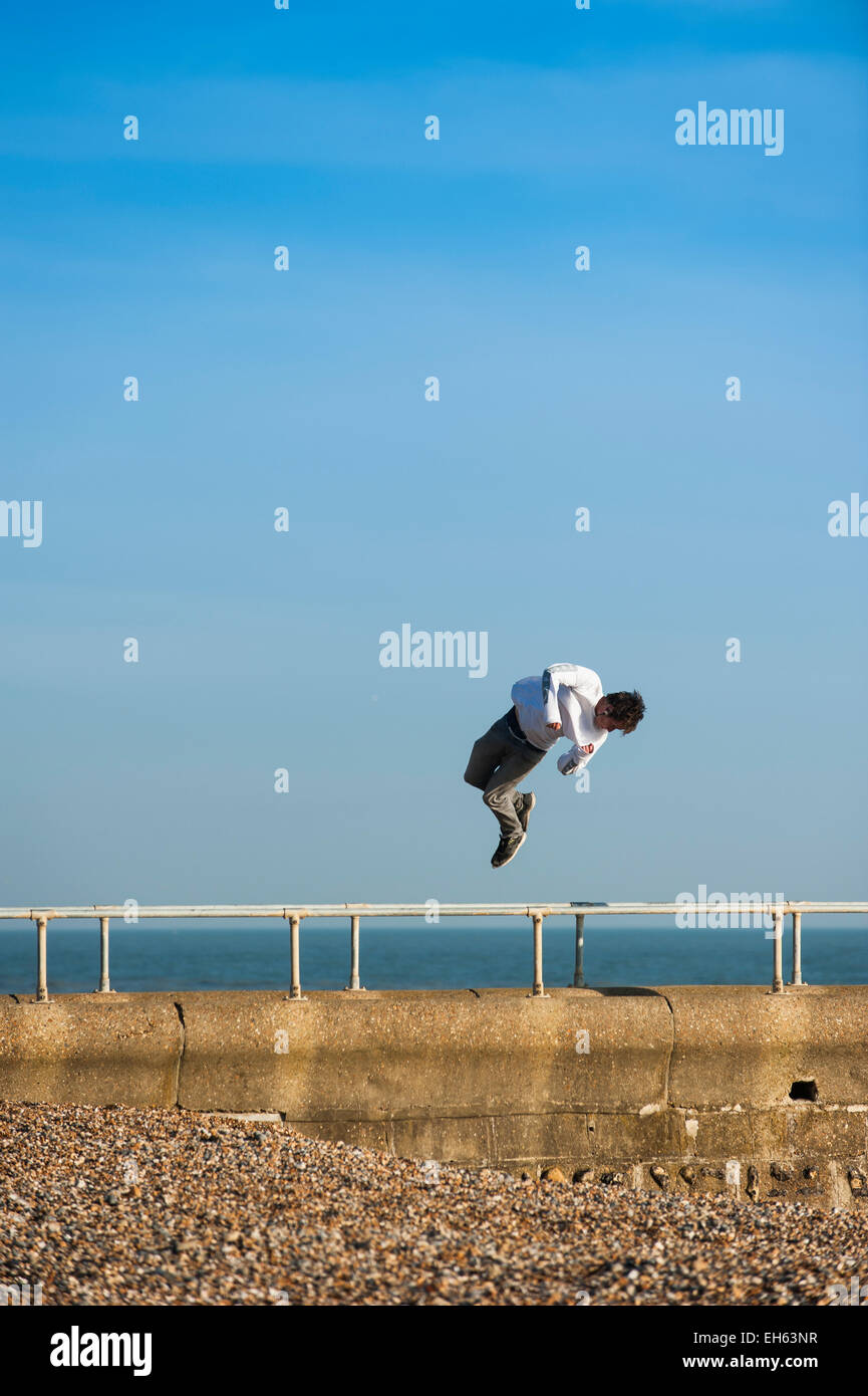 Un giovane uomo di torsioni in aria mentre un gruppo di giovani che stanno facendo la maggior parte della soleggiata primavera meteo praticare l'arte del parkour, free-jumping e facendo salti mortali, off un pontile sul lungomare di Brighton con il dis-usato Molo Ovest in background. Brighton, East Sussex, Inghilterra, Regno Unito. Foto Stock