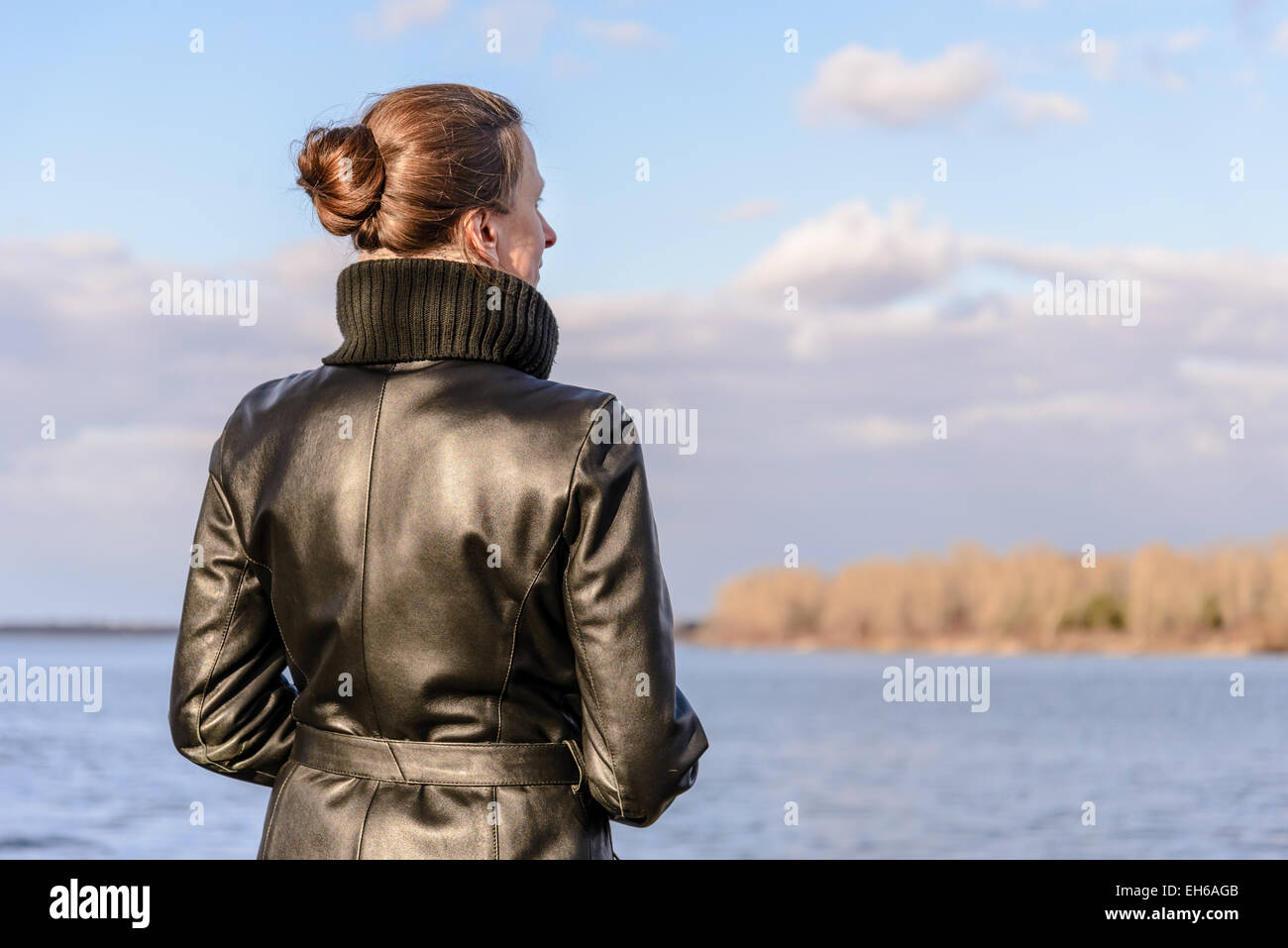 Una donna con un chignon e un nero Cappotto di pelle è guardare il paesaggio vicino al lago o fiume durante una giornata di sole con Foto Stock