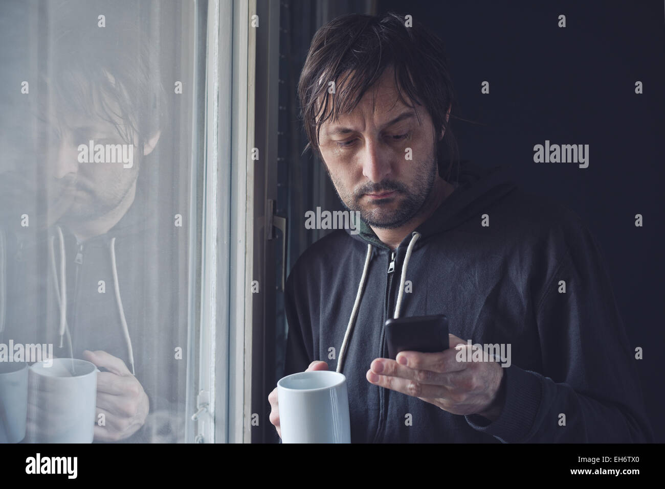 Uomo adulto a bere caffè e la lettura di un messaggio di testo o SMS sul cellulare in mattinata dalla finestra. Messa a fuoco selettiva con bassa Foto Stock