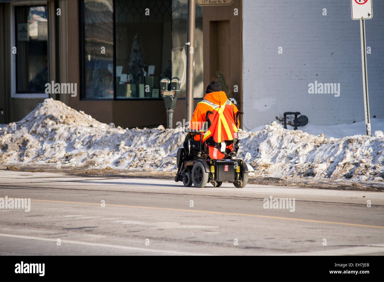 Uomo in sedia elettrica scooter riding down road accanto a snowbanks Foto Stock