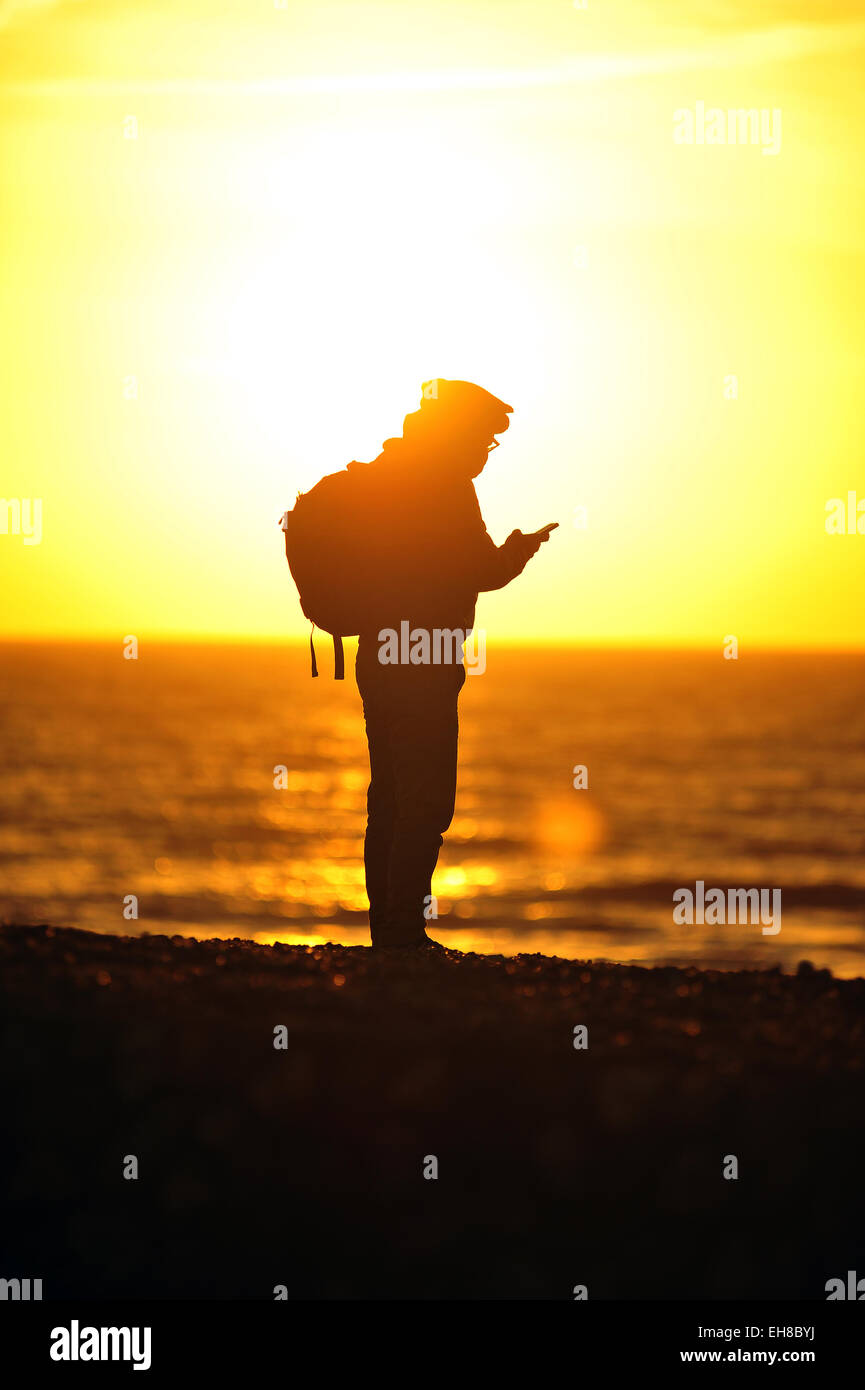 Un uomo che guarda il suo telefono si stagliano contro il sole sulla spiaggia di Brighton nel Regno Unito. Foto Stock