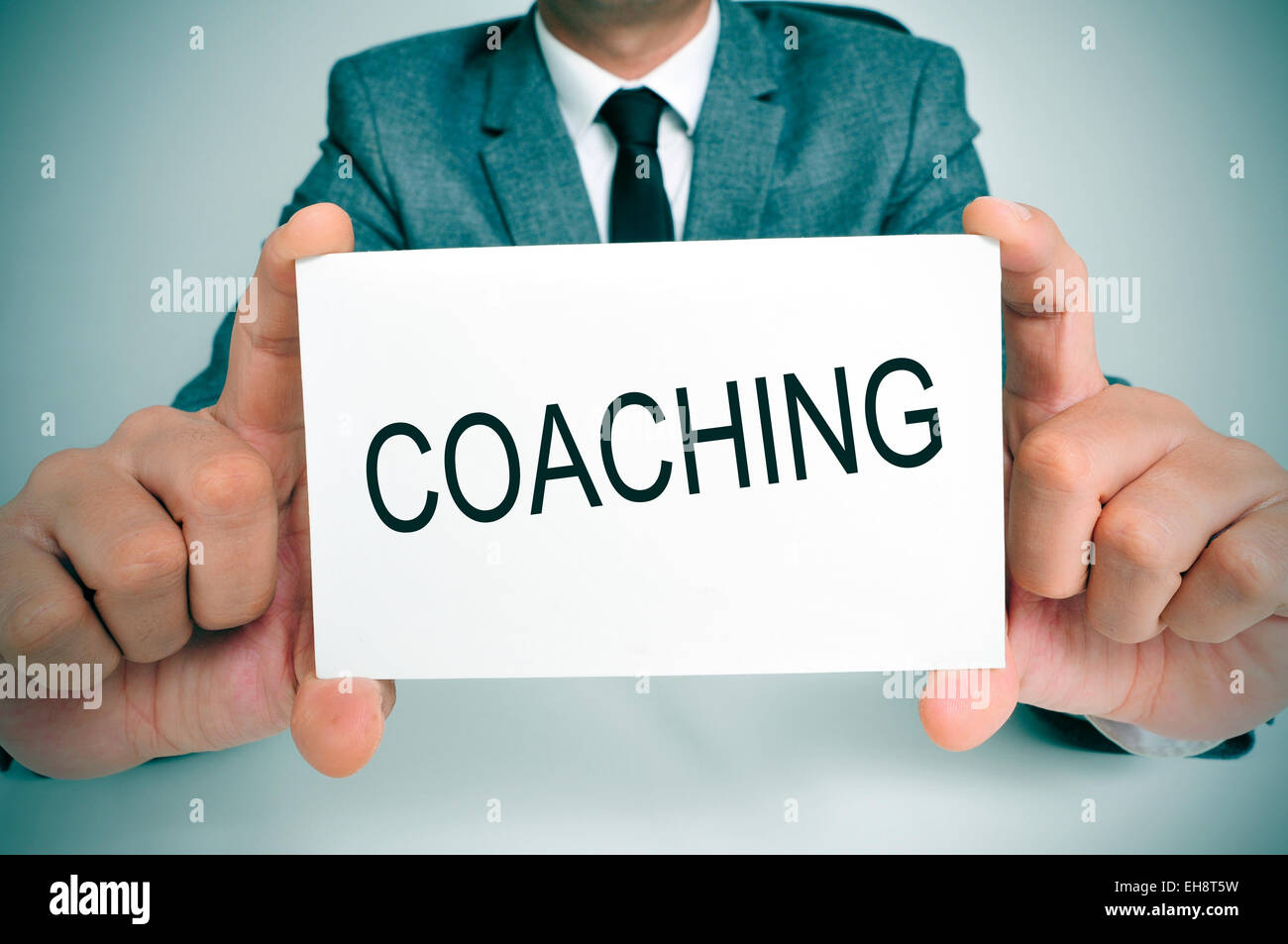 Un giovane uomo in tuta seduto alla sua scrivania in ufficio mostra un cartello con scritto il testo scritto di coaching in esso Foto Stock