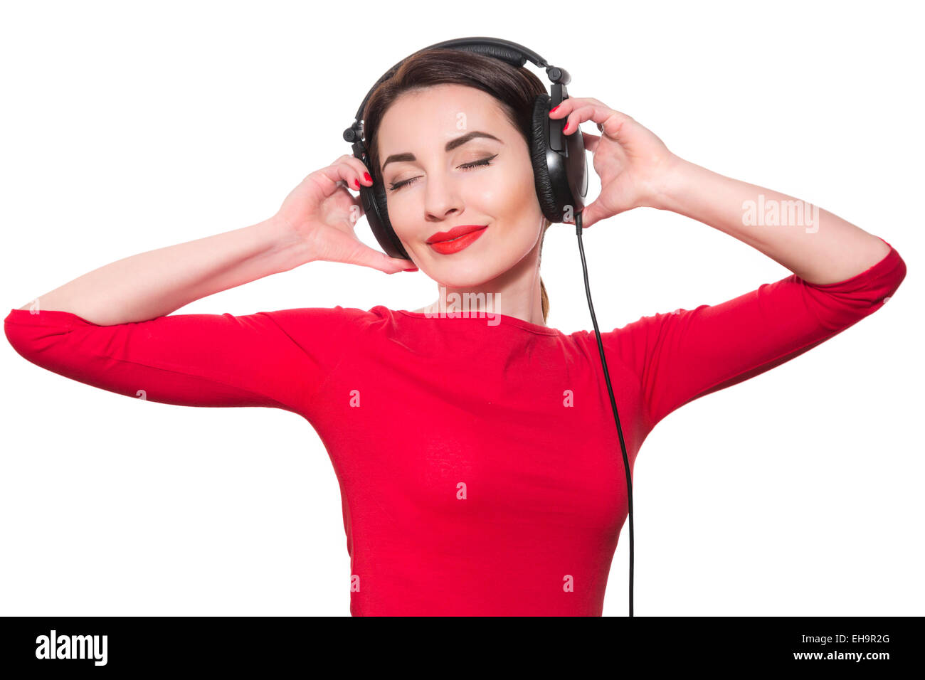 Attraente giovane donna adulta in rosso vestiti ascoltando la musica toccando grandi cuffie con gli occhi chiusi isolato su bianco ba Foto Stock