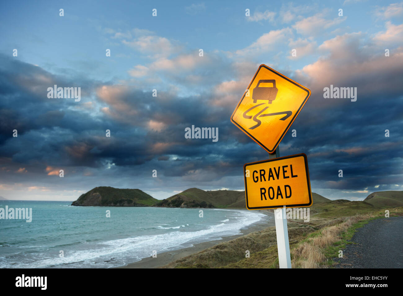 Attenzione (strada di ghiaia) segno sulla strada per mare contro il cielo nuvoloso al tramonto. Foto Stock