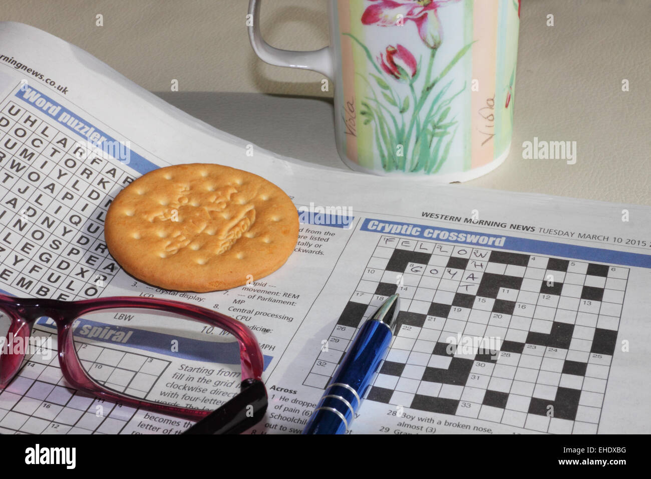 Un giornale aperto alla pagina dei cruciverba con gli occhiali da lettura, una penna, una tazza e biscotti. Foto Stock