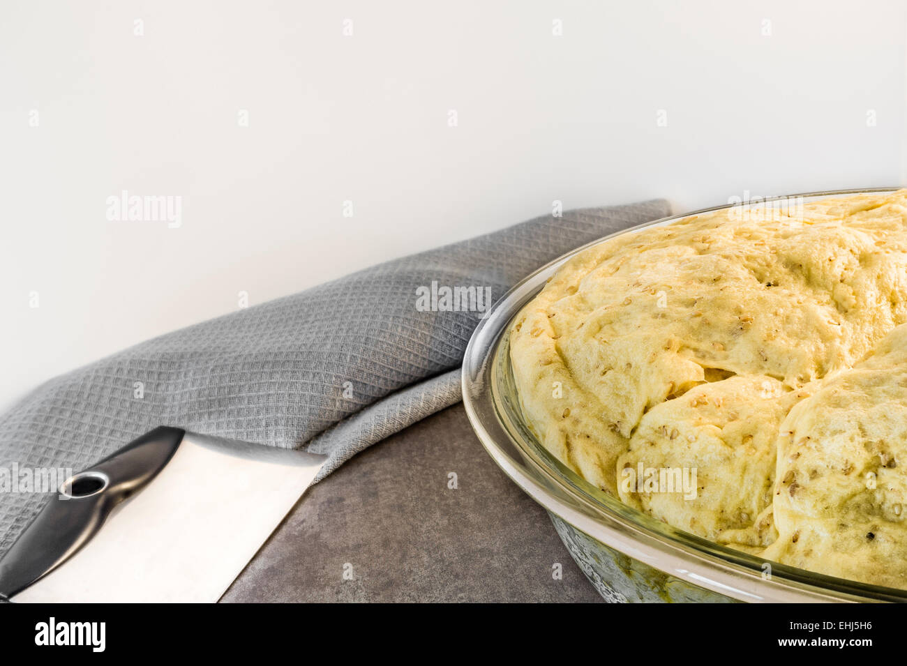 Un close-up di una ciotola di aumento di pasta di pane, un acciaio inossidabile raschiatore di pasticceria, su una superficie di cemento con un grigio asciugamano da cucina. Foto Stock