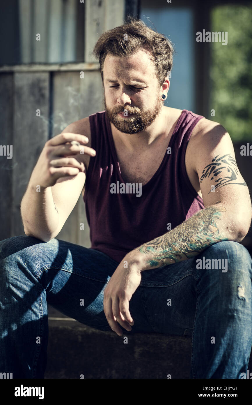 Un giovane uomo seduto al di fuori di una casa abbandonata e fumo di sigarette. Foto Stock