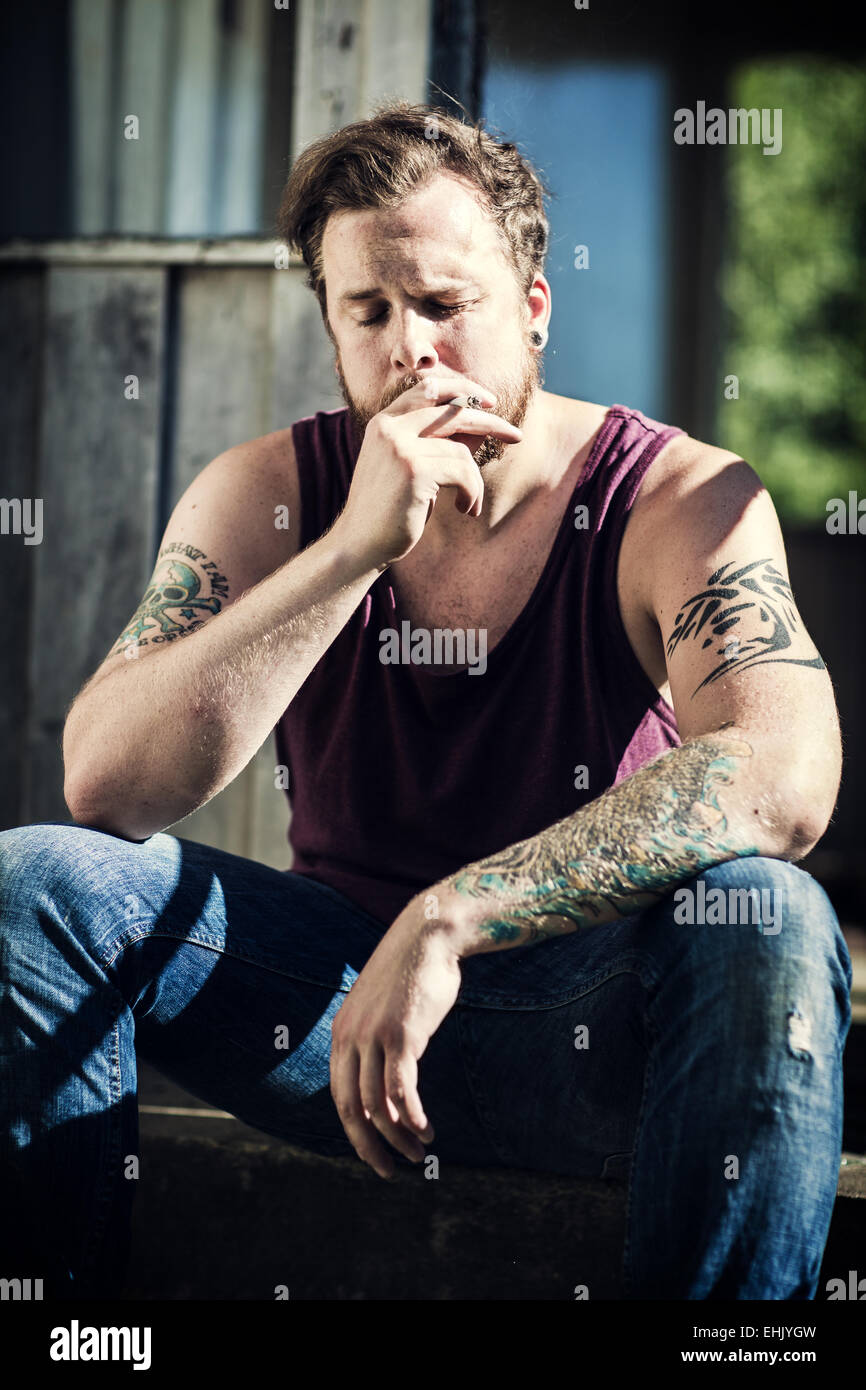 Un giovane uomo seduto fuori casa abbandonata e fumo di sigarette. Foto Stock