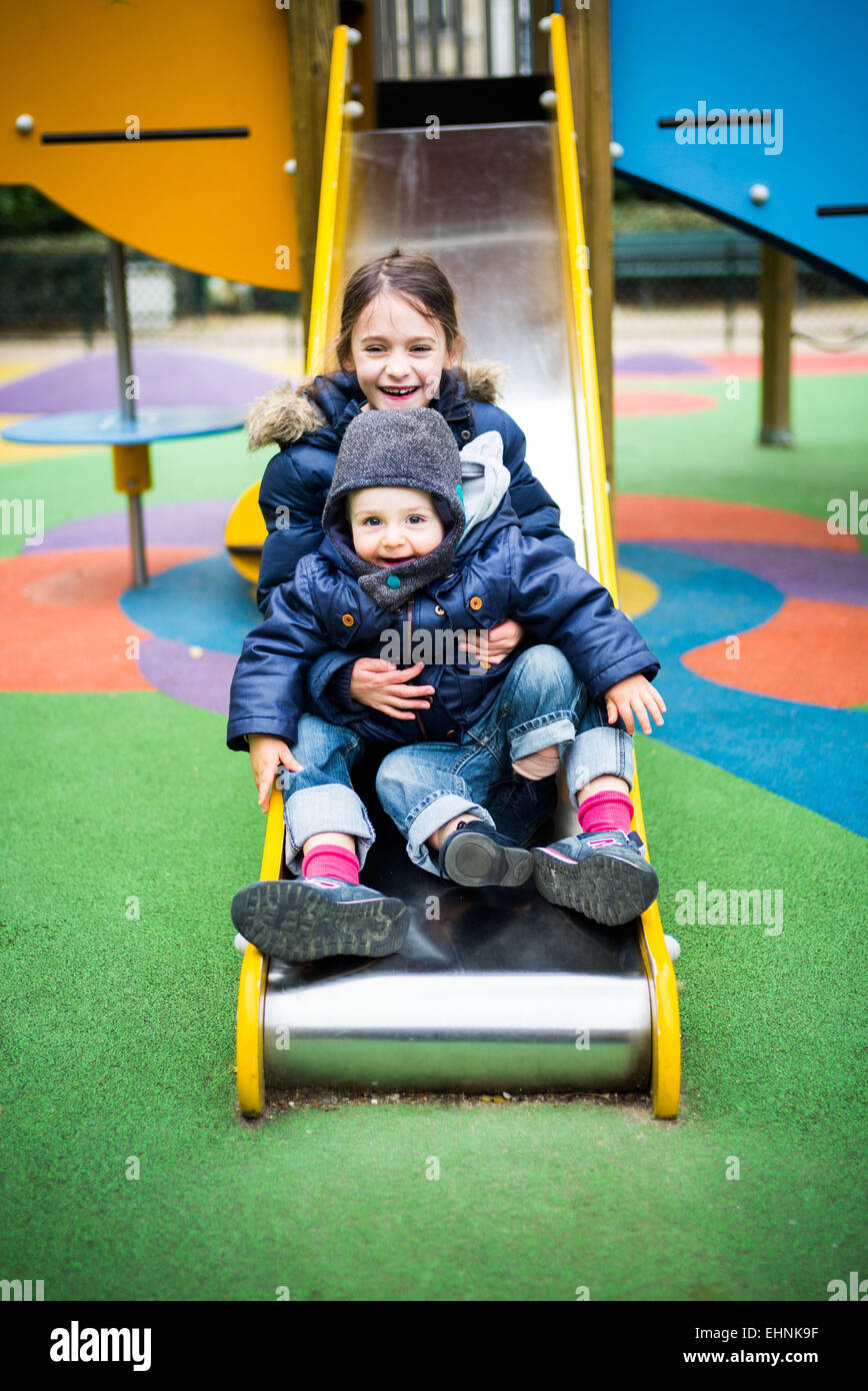 7 anno-vecchia ragazza e 18 mese-vecchio baby boy su una diapositiva in un parco giochi. Foto Stock
