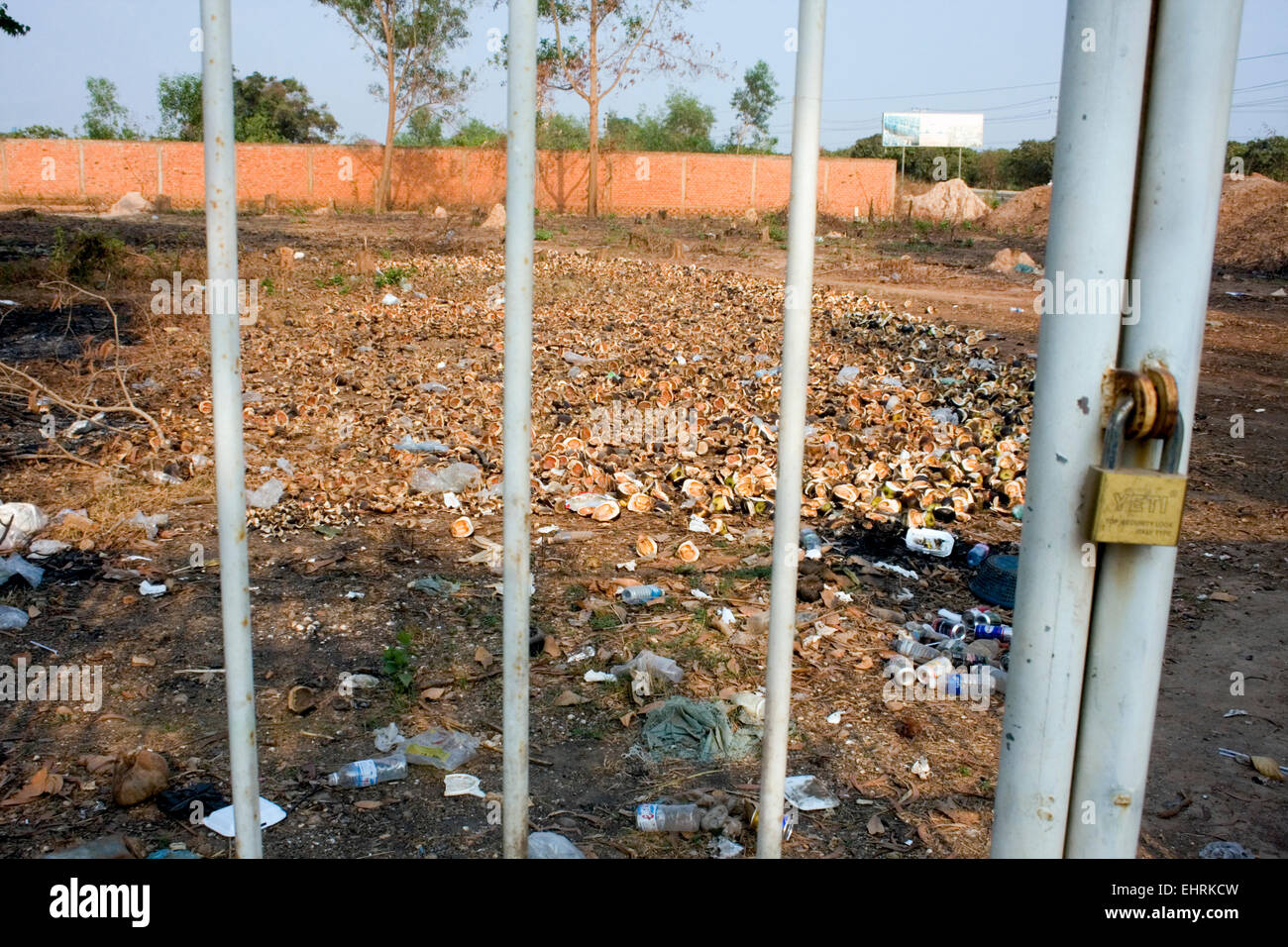 Aprire un campo disseminato di spazzatura e gusci di noce di cocco è visto attraverso un cancello chiuso con lucchetto in Skun, Kampong Cham Provincia, in Cambogia. Foto Stock