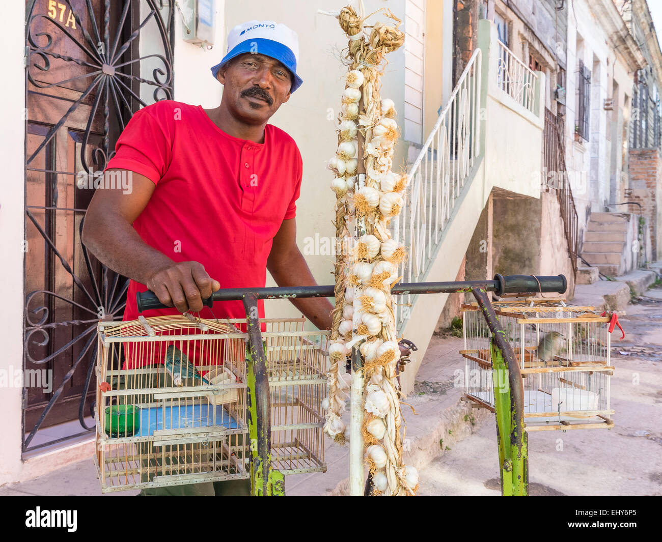 Un cubano di uomo di mezza età vende aglio e uccelli in gabbie dal suo carro ha ruote sulle strade di Regla, Cuba. Foto Stock