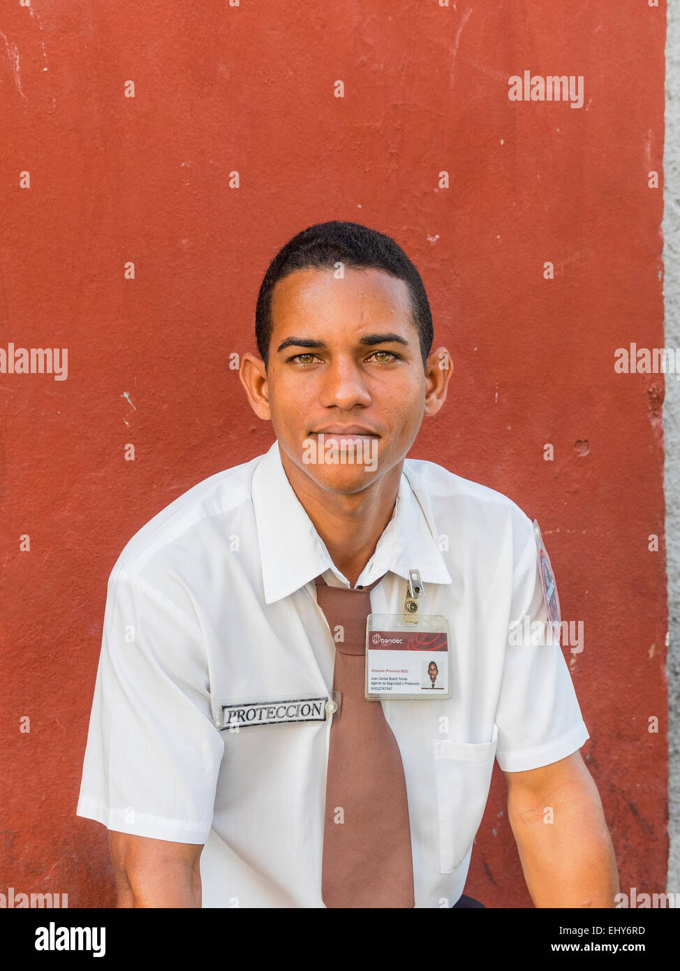 Giovani adulti cubano guardia di sicurezza si appoggia contro una parete rossa che indossa una camicia bianca e cravatta marrone e il suo distintivo di identificazione. Foto Stock