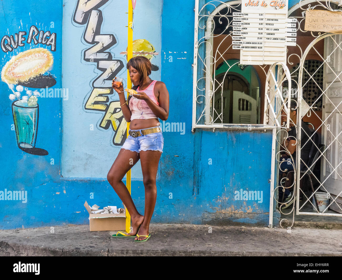 Una donna afro-cubane di mangiare la pizza davanti al cibo e di stallo in piedi di fronte a un profondo blu del muro con un segno dipinto annuncio Foto Stock