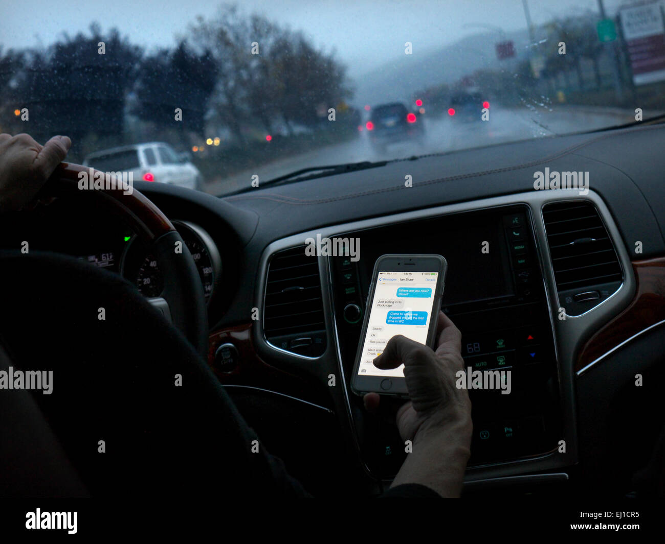 In auto texting driver femminile in l/h guidare il veicolo nascostamente texting smartphone durante la guida sul bagnato trafficata autostrada al crepuscolo Foto Stock