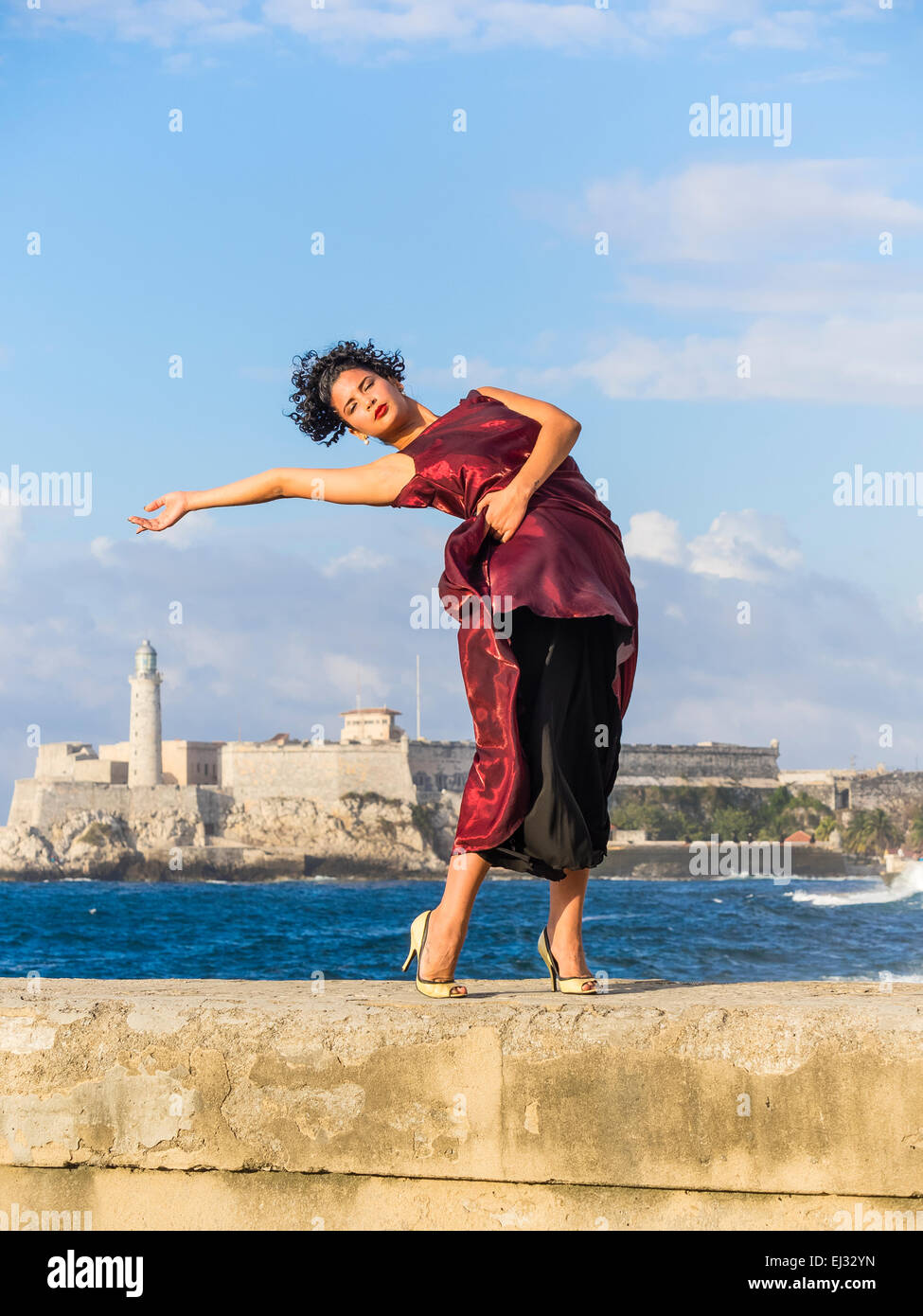 26 anno cubano ispanica modello femminile in posa di maroon abiti sul Malecón con il muro di Morro storico castello in background. Foto Stock