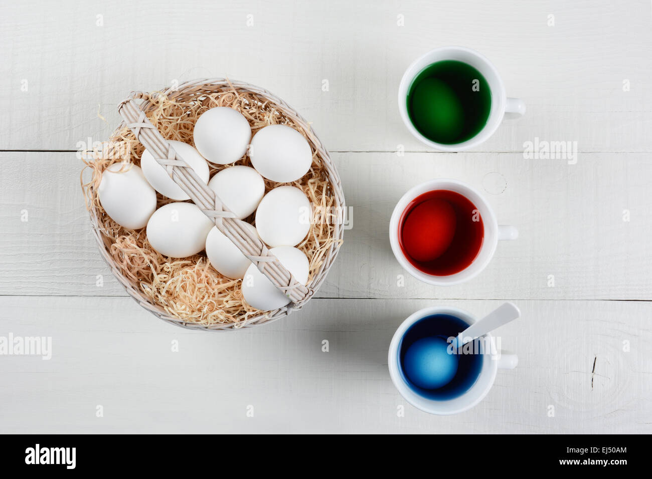 Alta angolazione di un cesto pieno di uova per essere tinti per la Pasqua. Tre tazze di colorante con uova ammollo sono accanto ai cestelli di u Foto Stock