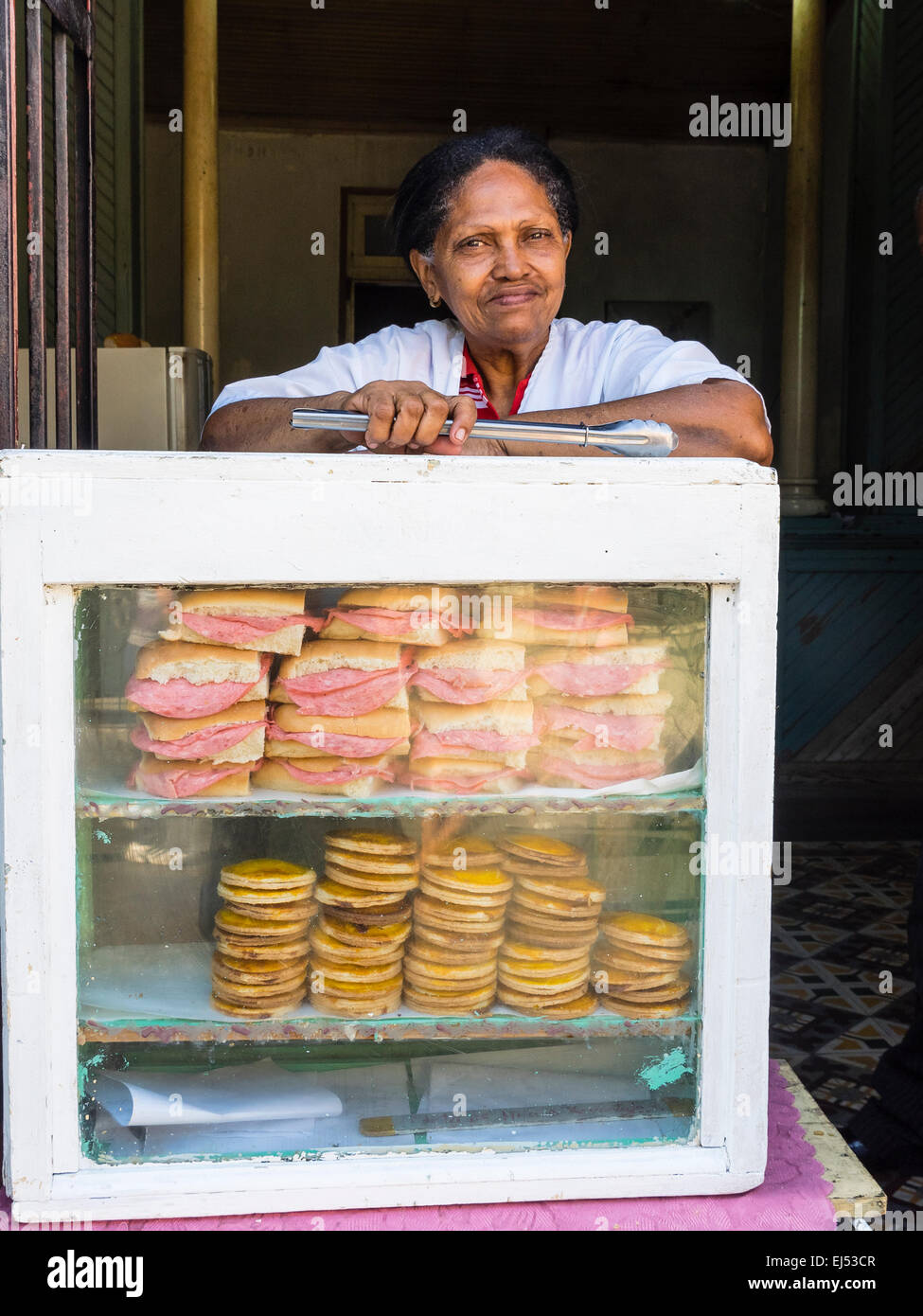 Un vecchio afro-cubane sandwich femmina venditore si appoggia sul suo caso di visualizzazione azienda panini e altri alimenti mantenendo le tenaglie e l Foto Stock