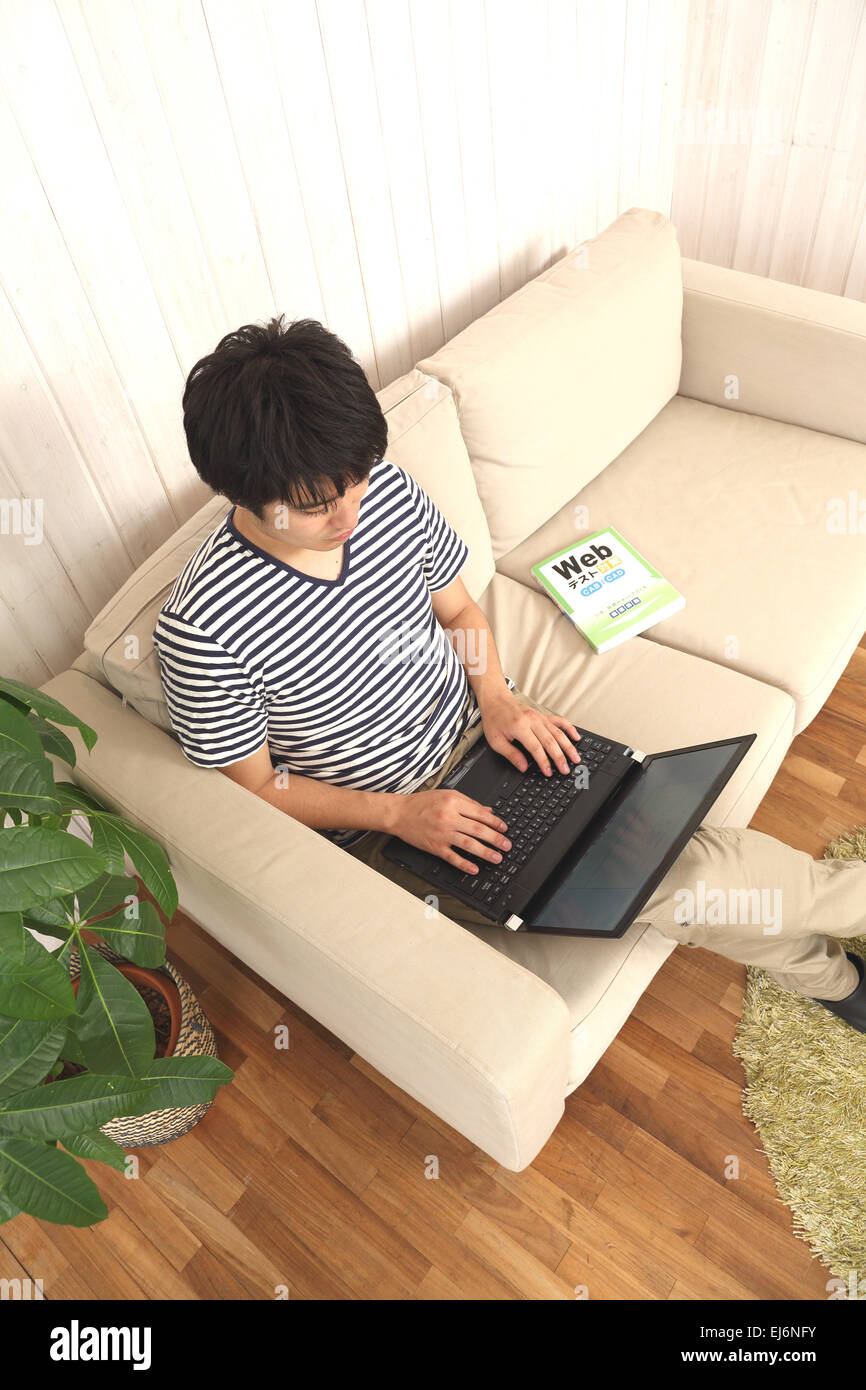 Un giovane giapponese uomo che lavora presso il suo computer portatile sul divano Foto Stock