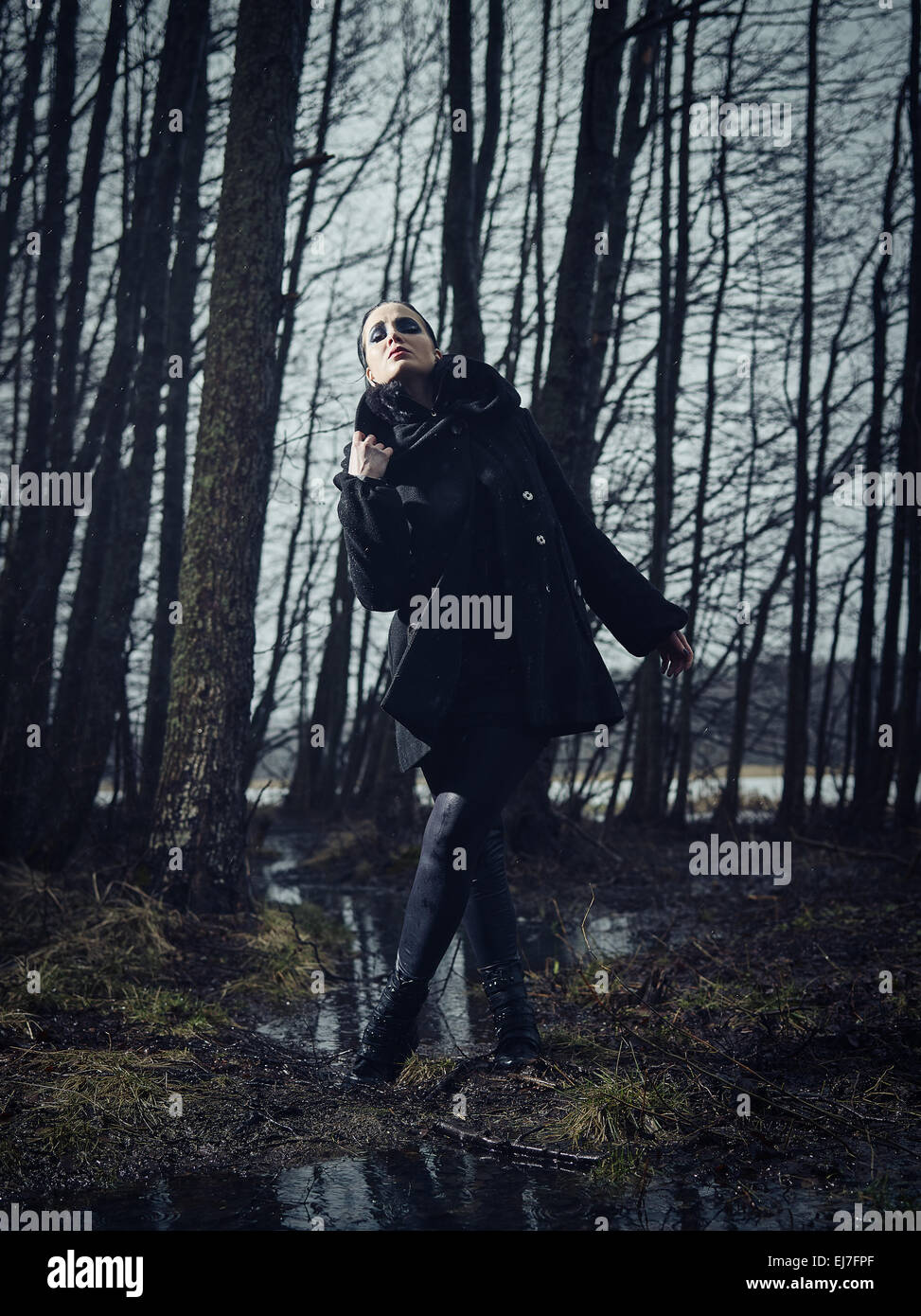 Moda donna che indossa un cappotto invernale e lei in piedi in una cupa foresta, freddo pioggia, Croce trasformati a piena lunghezza immagine Foto Stock