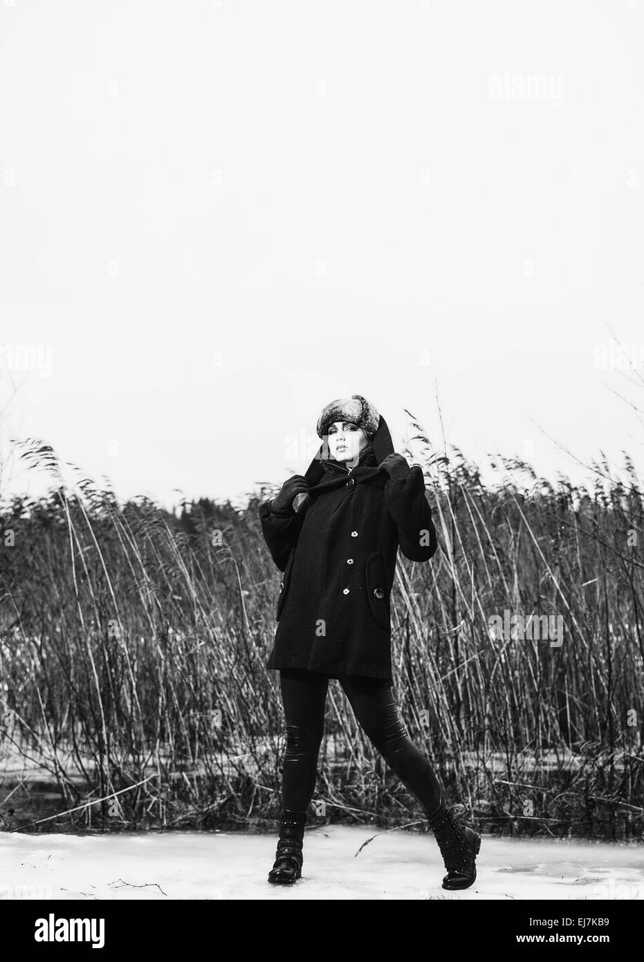 Moda donna che indossa un cappotto invernale e cappuccio di pelliccia e lei in posa davanti dei canneti, freddo pioggia a piena lunghezza nero e bianco Foto Stock