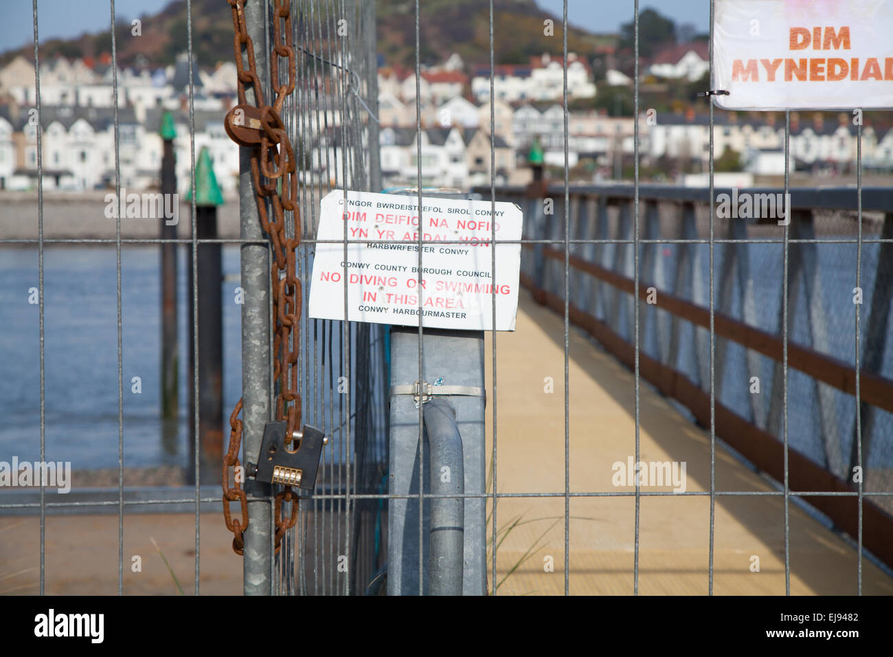 Maglia di filo recinzioni e cartelli di avvertimento attraverso un pontile in disuso a Conwy Harbour, il Galles del Nord a bassa marea e con Deganwy dietro Foto Stock