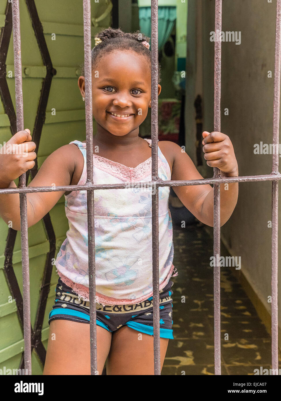 Una giovane ragazza afro-cubane trattiene le barre della porta di sicurezza a casa sua con entrambe le mani e si affaccia sulla strada e sorrisi. Foto Stock