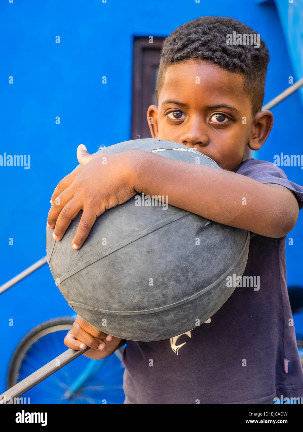 Un ragazzo afro-cubane tenendo un vecchio battere fino a basket in un braccio e nella parte anteriore del suo corpo rivolta in avanti contro il muro di blu. Foto Stock