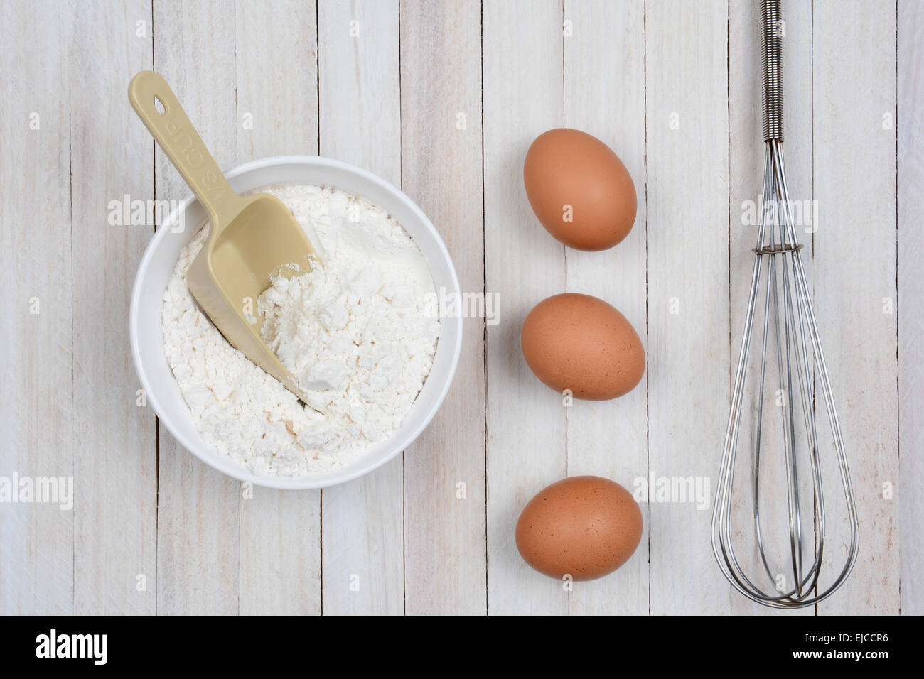 Immagine aerea di una tazza di farina, 3 uova di colore marrone e una frusta di metallo su un legno rustico tavolo da cucina. Foto Stock