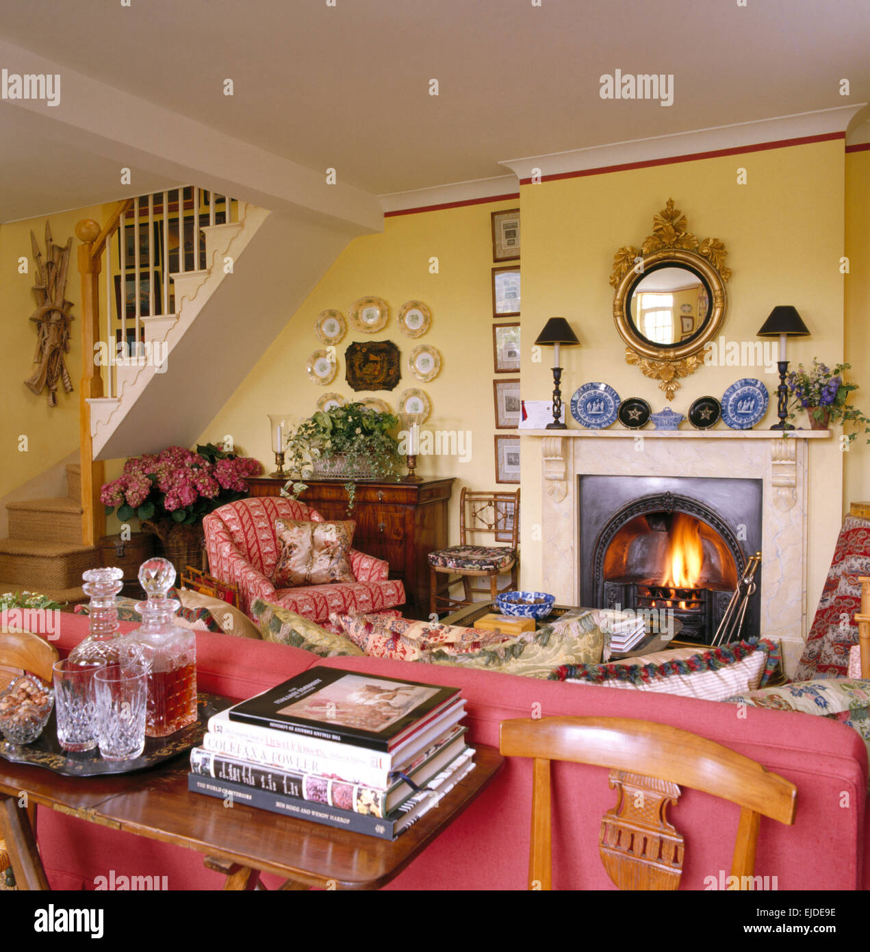 Tagliare decantatori di vetro sul tavolo dietro il divano rosso in giallo cottage salotto con uno specchio antico camino sopra Foto Stock