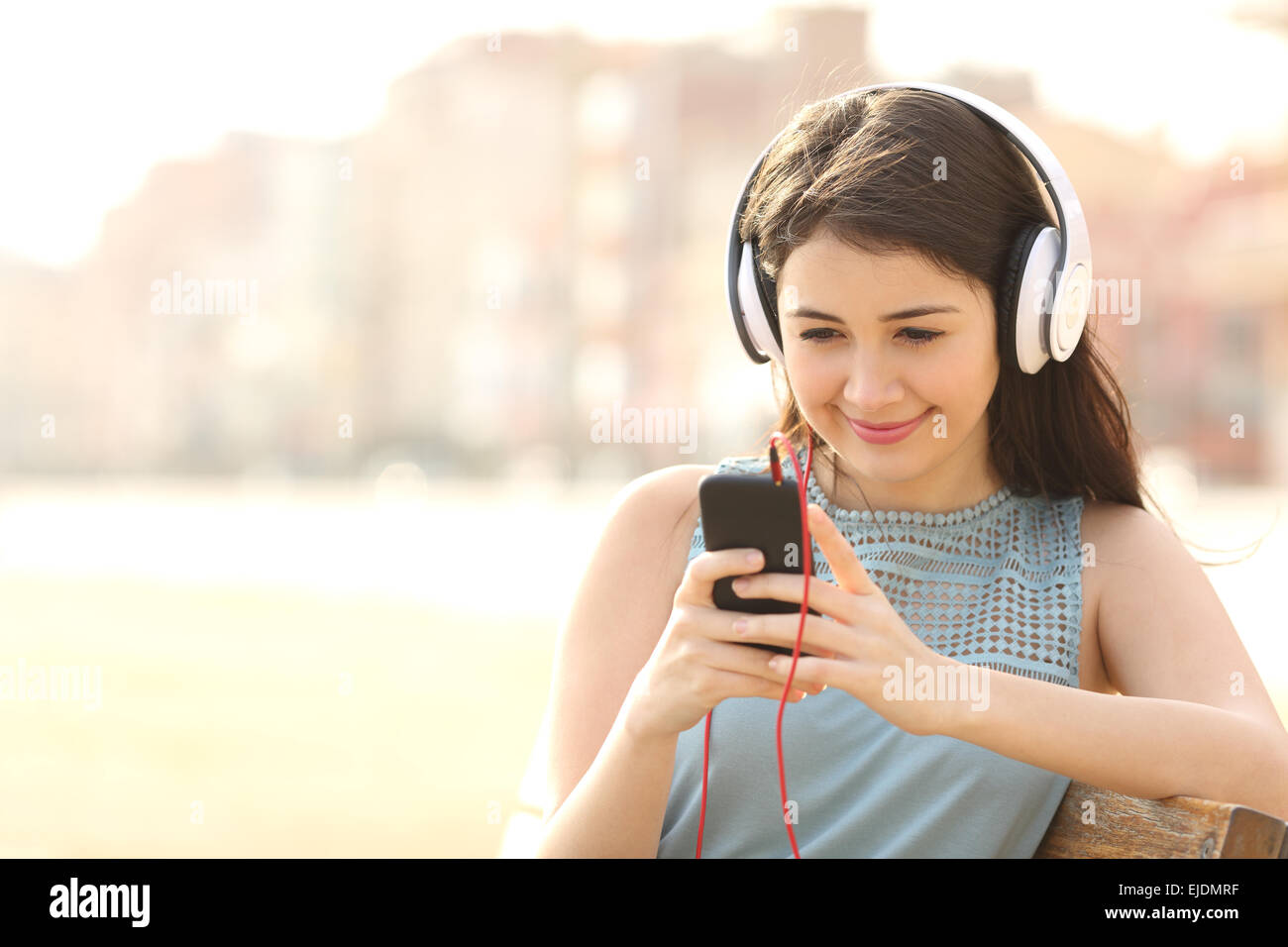 Ragazza ascolto di musica con le cuffie da uno smart phone seduto su una panchina in un parco Foto Stock