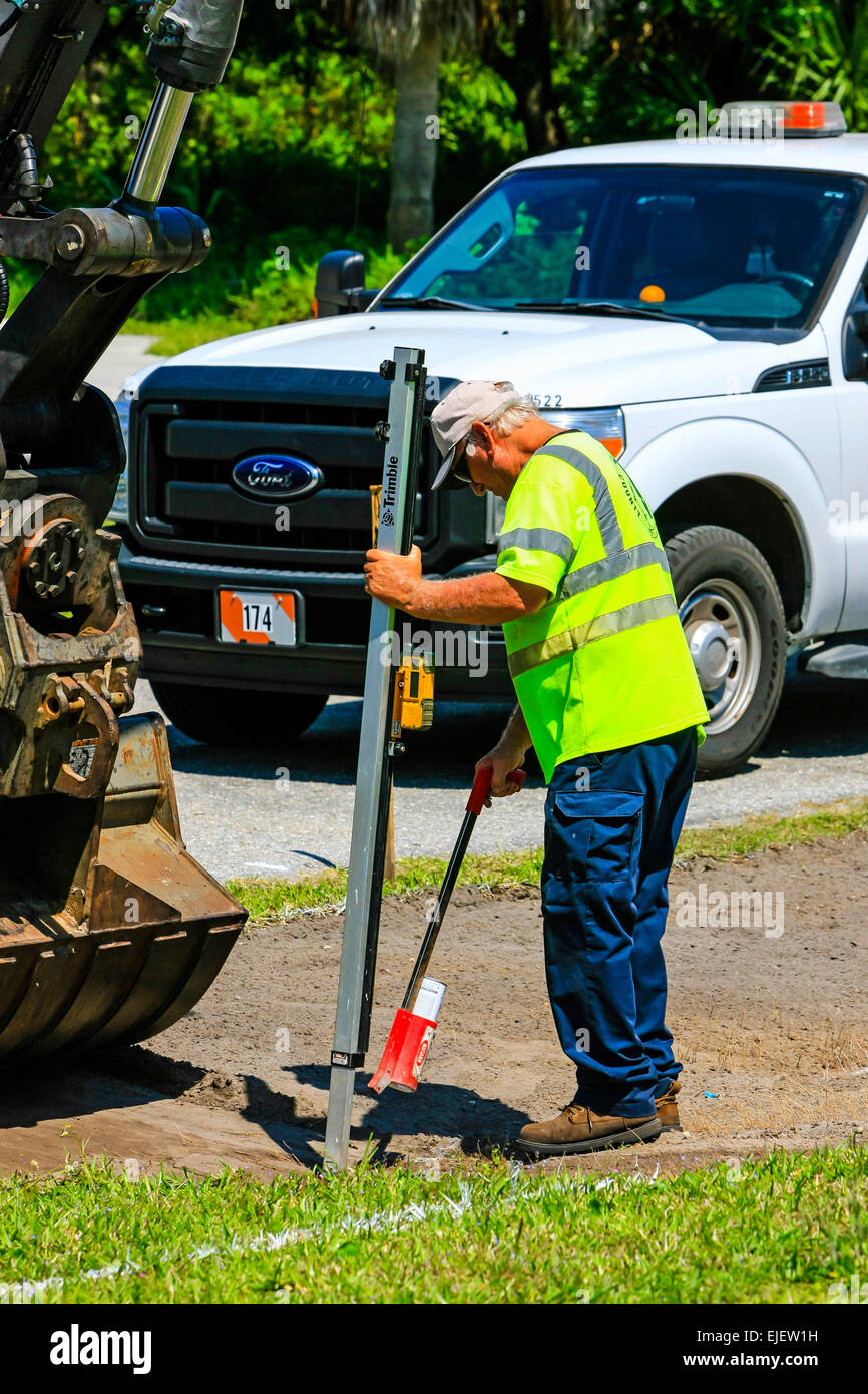 Autostrada lavoratore di manutenzione utilizzando una profondità digitale guage come il retroescavatore rimuove il terreno come parte dell'acqua piovana soak-lontano il drenaggio Foto Stock