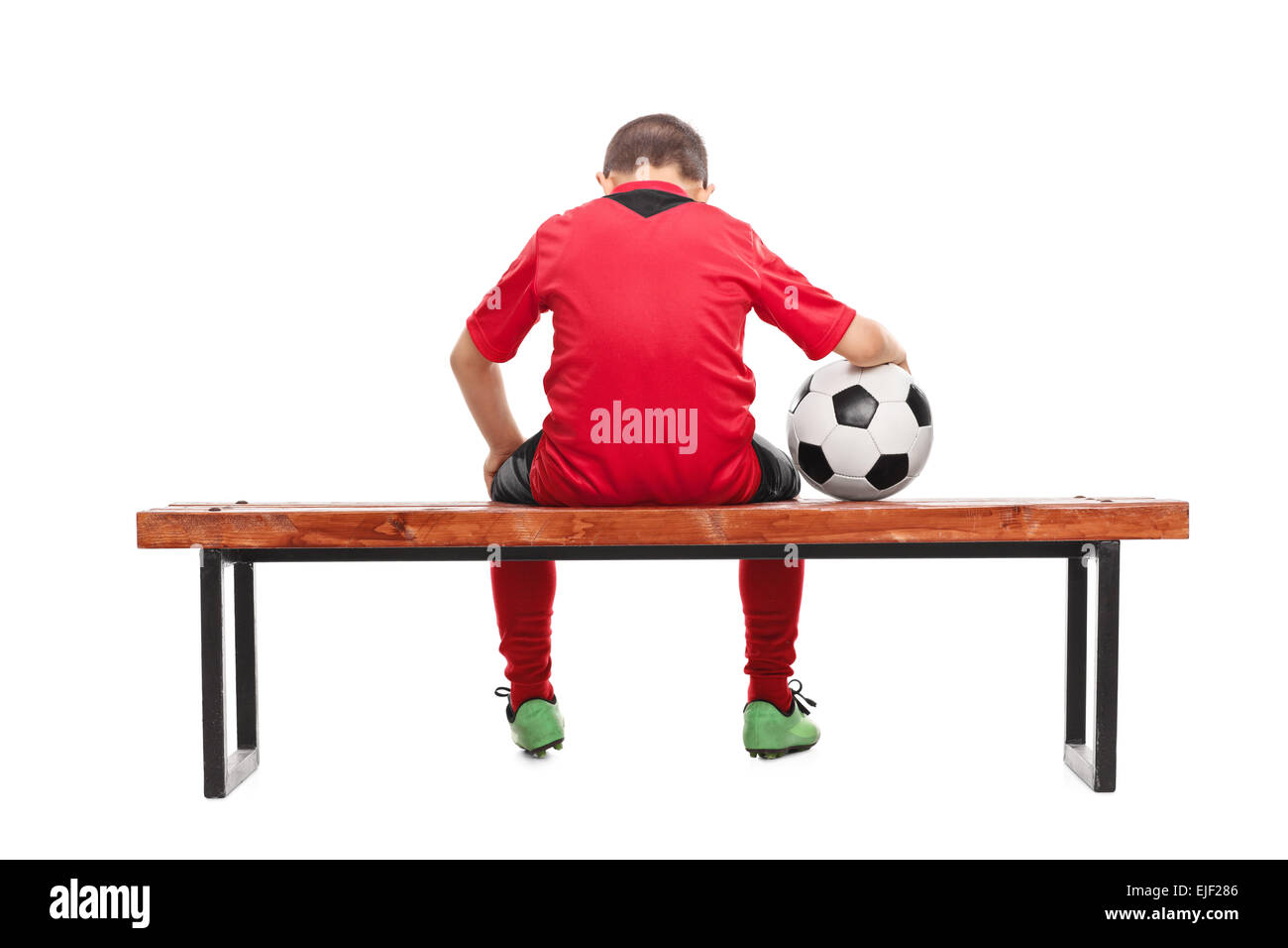 Vista posteriore studio shot di un triste little boy in rosso Soccer jersey seduto su una panchina e tenendo una palla isolati su sfondo bianco Foto Stock