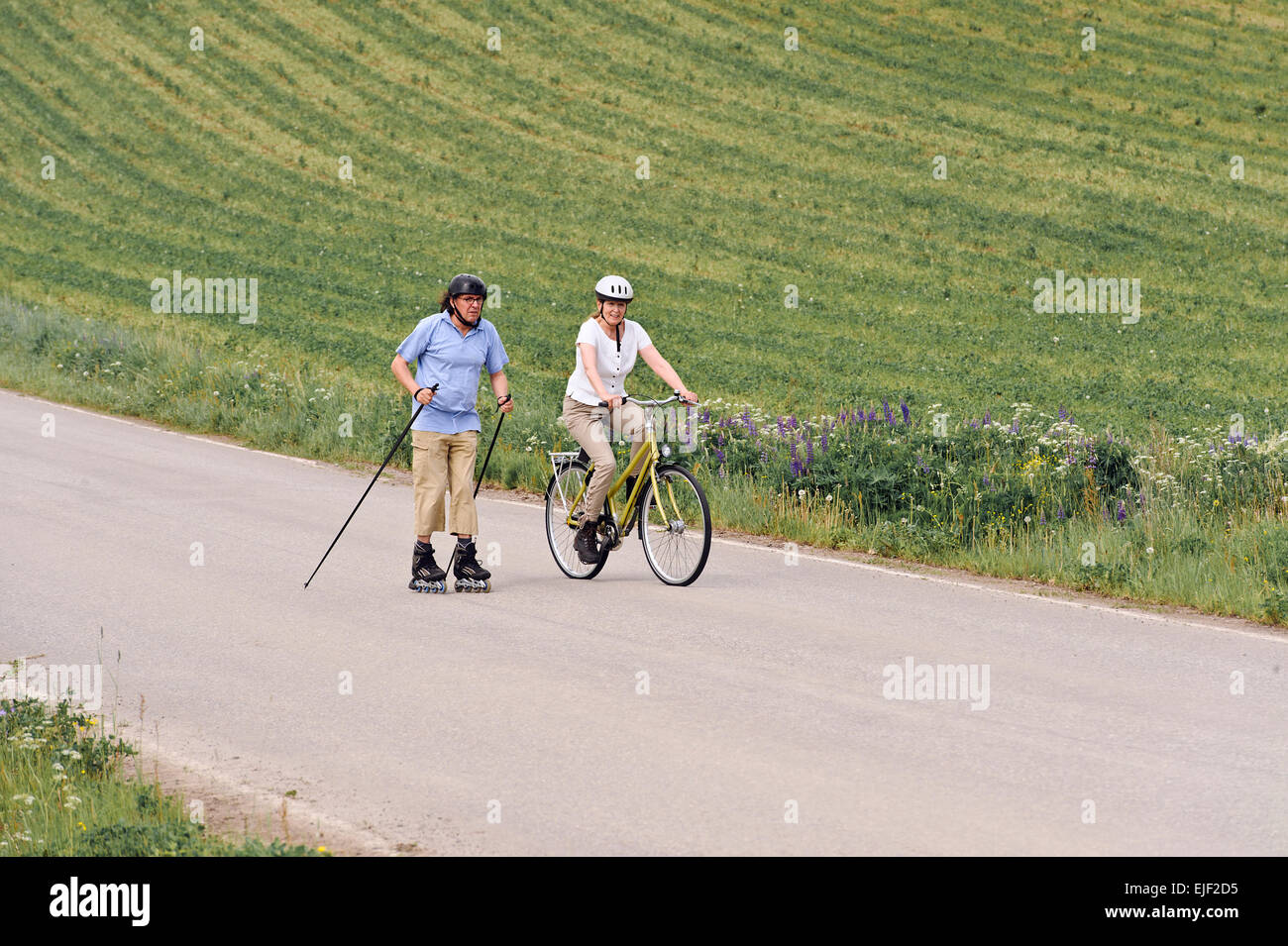 Coppia senior vigorosamente esercizio. L'uomo è nordico pattinaggio inline e la donna è in bicicletta. Si trova su una strada di campagna Foto Stock