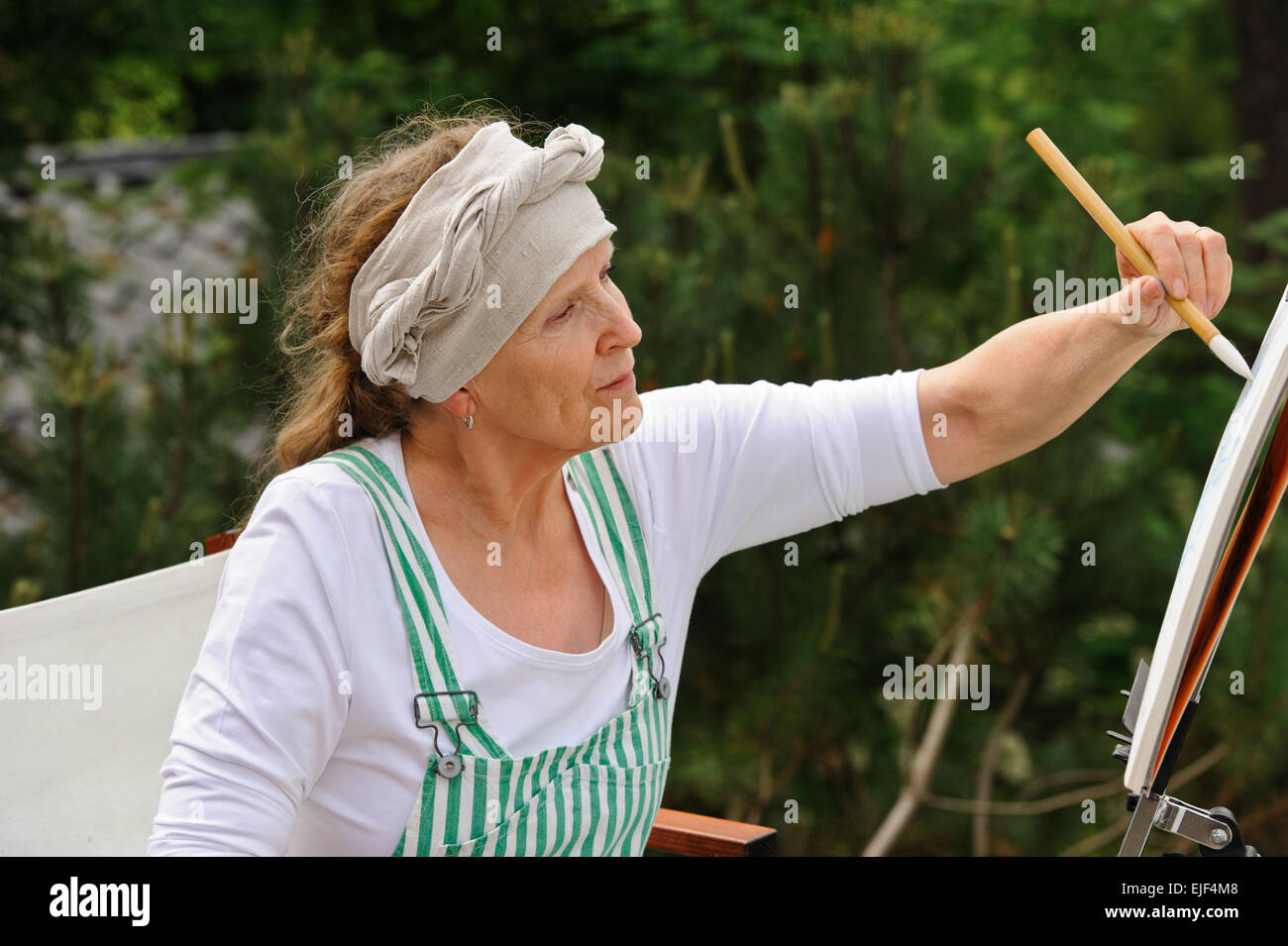 Senior donna pittura all'esterno. Si siede in una sedia di tela di fronte al cavalletto. Il giorno è coperto dando una luce morbida Foto Stock