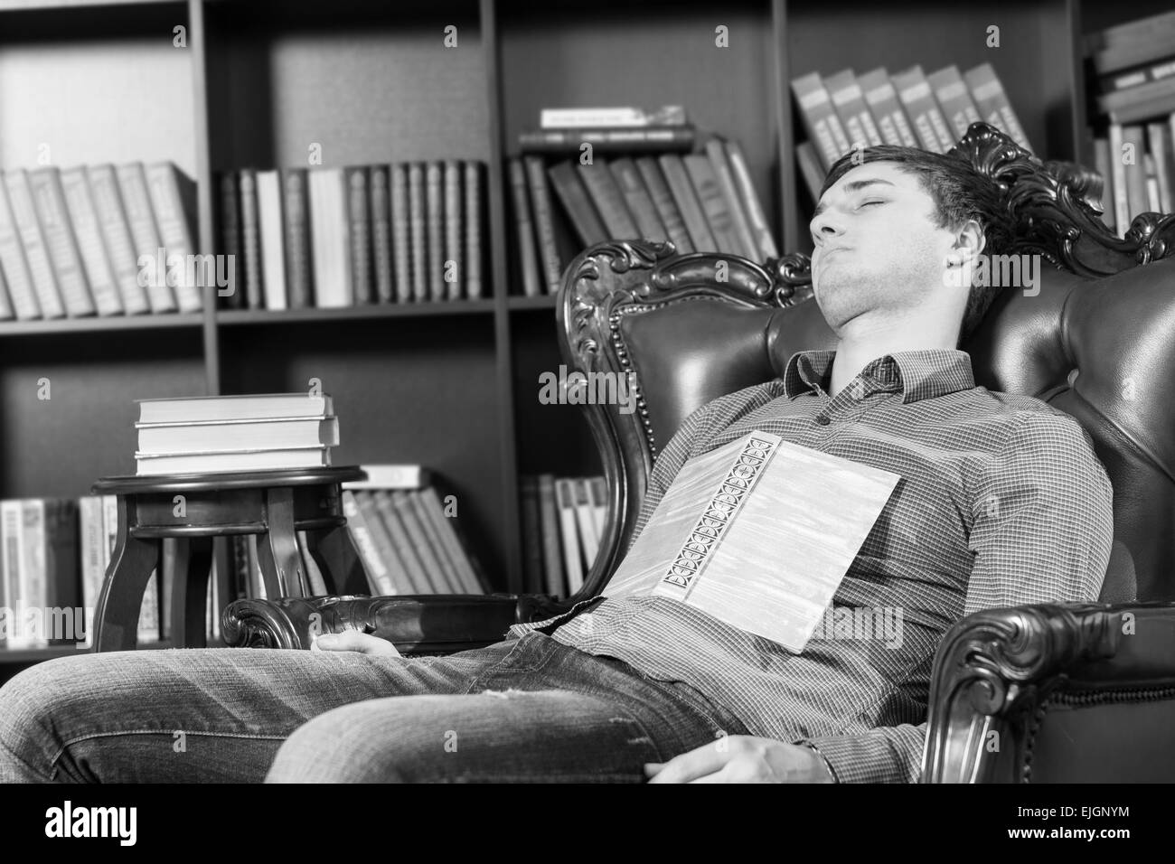 Close up stanco Giovane leggendo la letteratura in appoggio sulla sedia vicino gli scaffali con Libro sdraiato sul suo petto, catturato in monocromatico.. Foto Stock