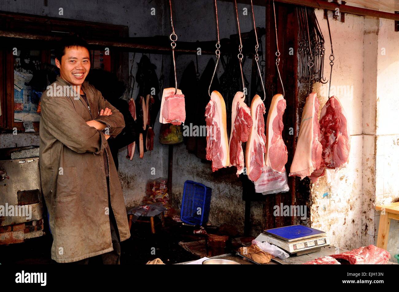 Pengzhou, Cina: Butcher in piedi nel suo negozio con vari tagli di carne di maiale appeso dal ferro ganci a base di carne Foto Stock