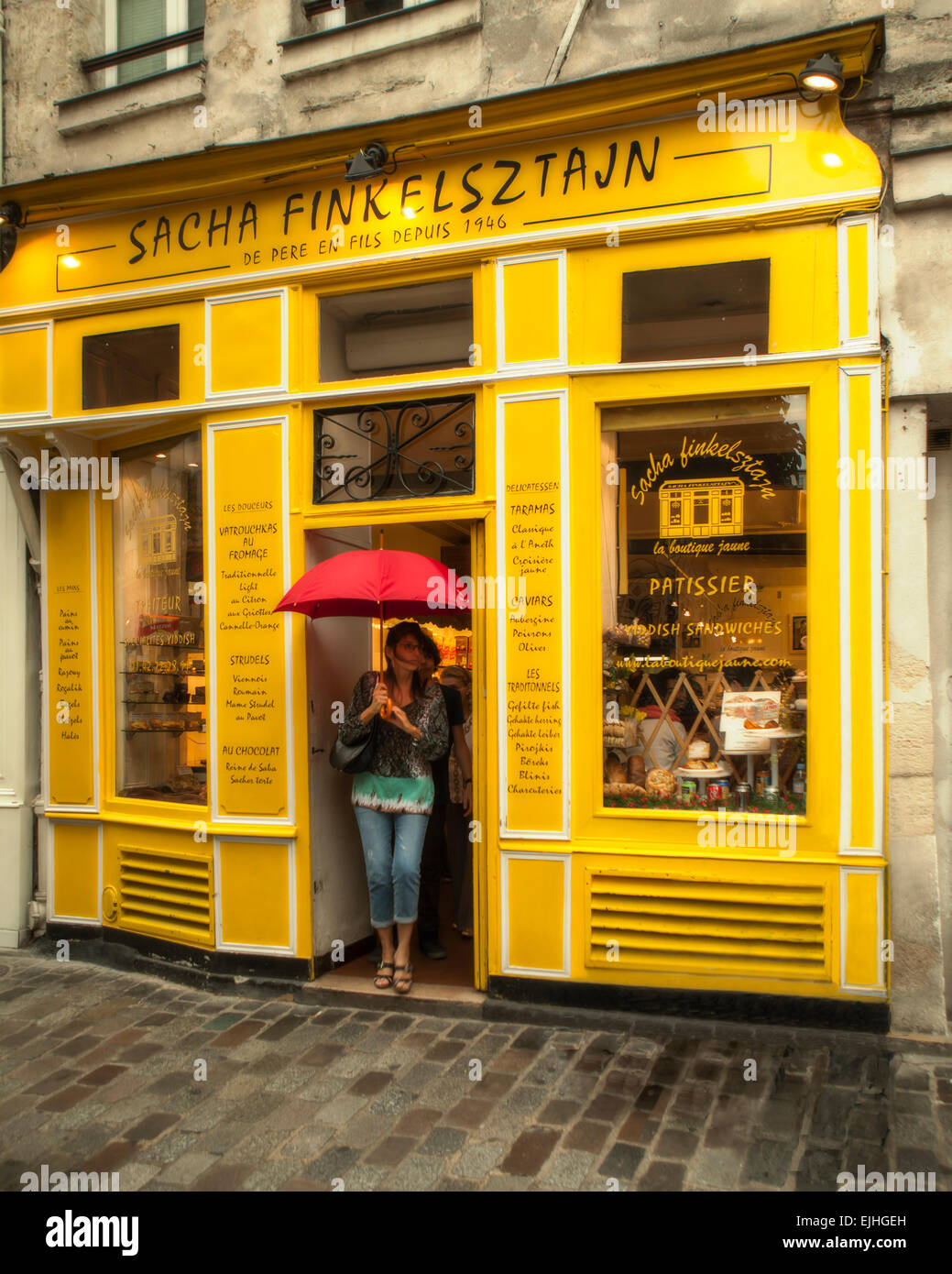 Donna con ombrello rosso in piedi presso la porta di colore giallo brillante pasticceria a Le Marais, Parigi, Francia Foto Stock