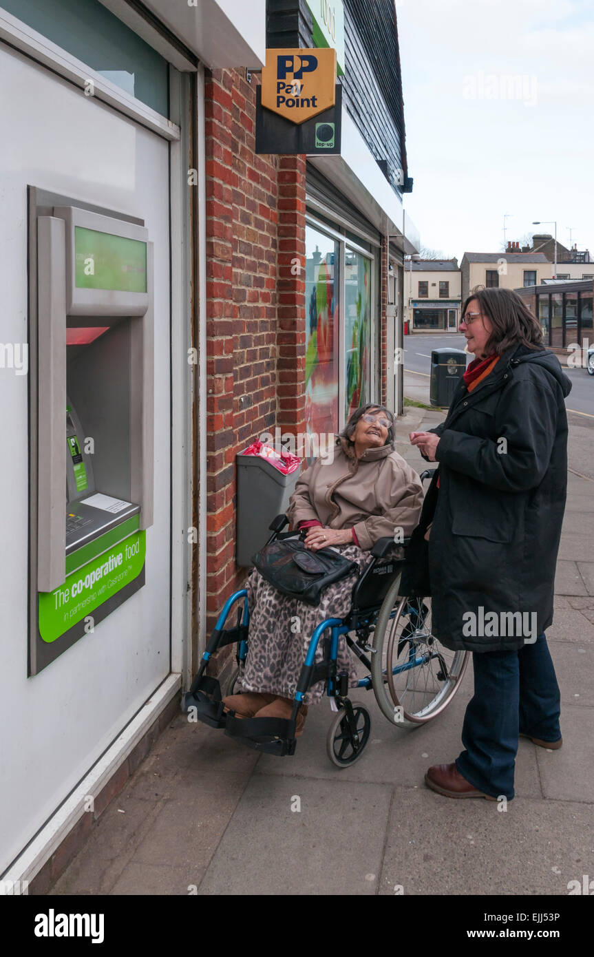 Un anziano di 90 anni lady in una sedia a rotelle è aiutato a ritirare i soldi da un bancomat ATM dal suo accompagnatore o assistente. Foto Stock