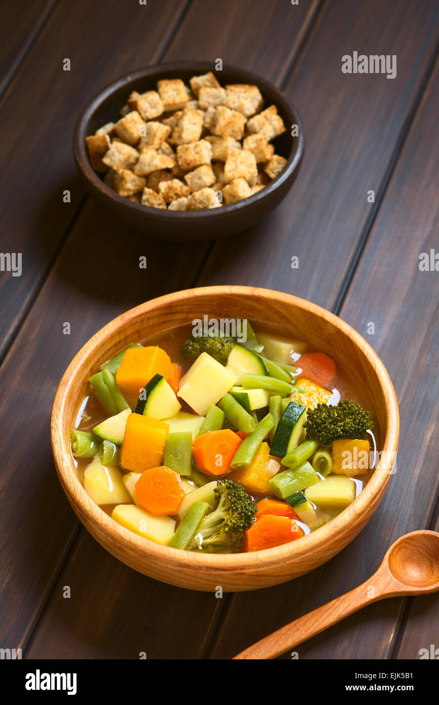 Ciotola di legno di origine vegetale zuppa di zucchine, verde fagiolo, carote, broccoli, patate e zucca con una piccola ciotola di crostini Foto Stock