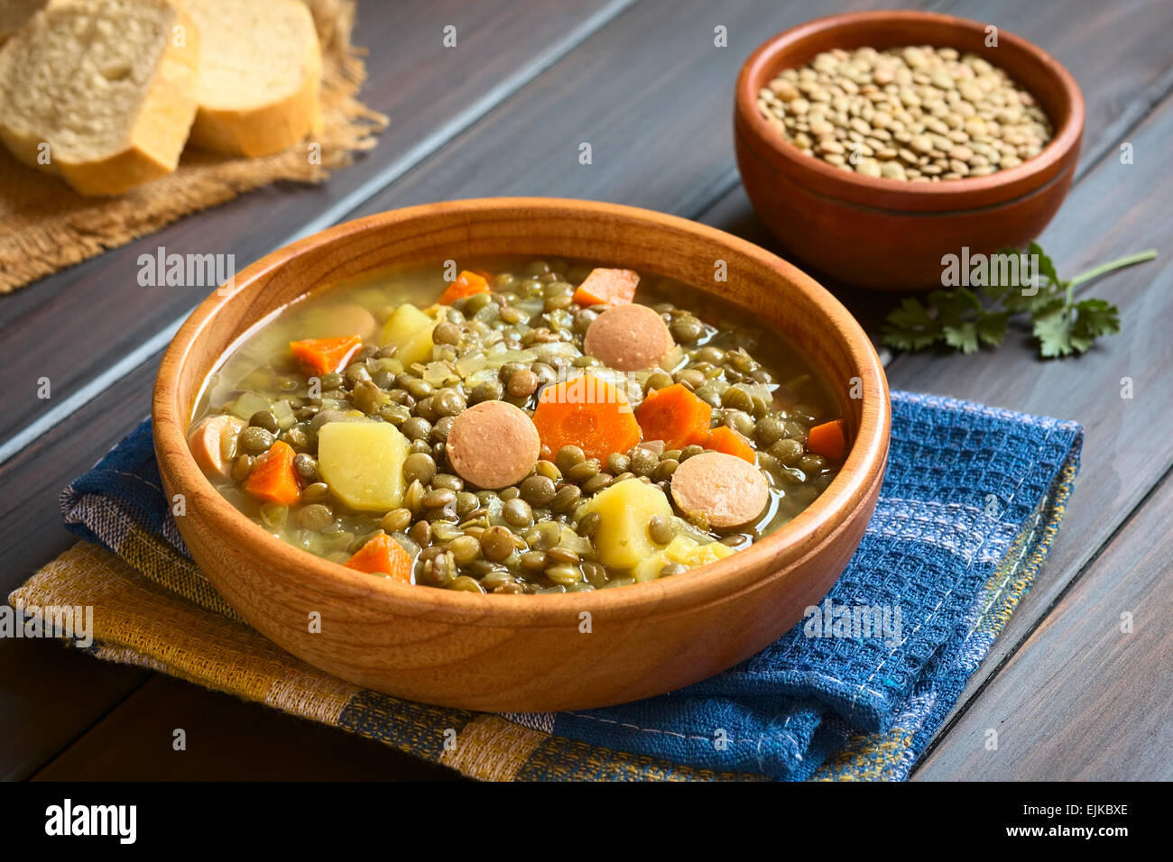 Ciotola di legno di zuppa di lenticchie fatta con la patata, la carota e la cipolla e le fette di salsiccia, con una piccola ciotola di lenticchie grezzo nella parte posteriore Foto Stock