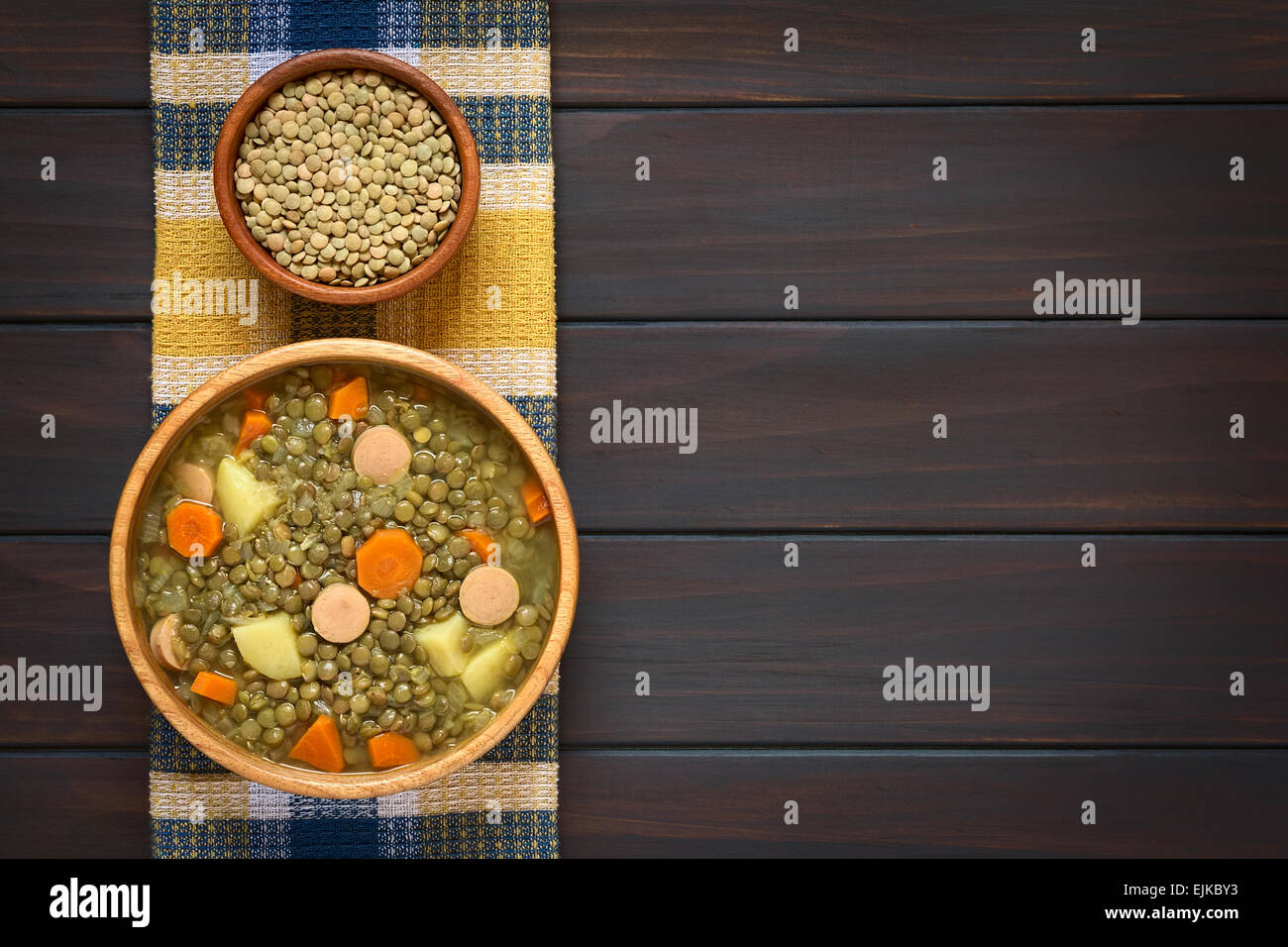 Scatto dall'alto di una ciotola di legno di zuppa di lenticchie fatta con la patata, la carota e la cipolla e le fette di salsiccia, con una piccola ciotola di materie lenticchie Foto Stock